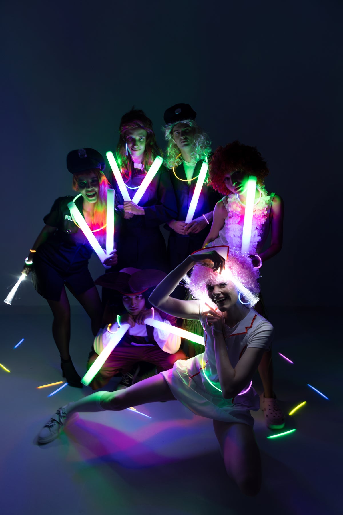 Five people posing together in a dark studio setting, each holding glowing neon light sticks that cast colorful reflections on the floor. The group is dressed in dark outfits with some wearing wigs, and the neon lights create a vibrant multicolor glow.
