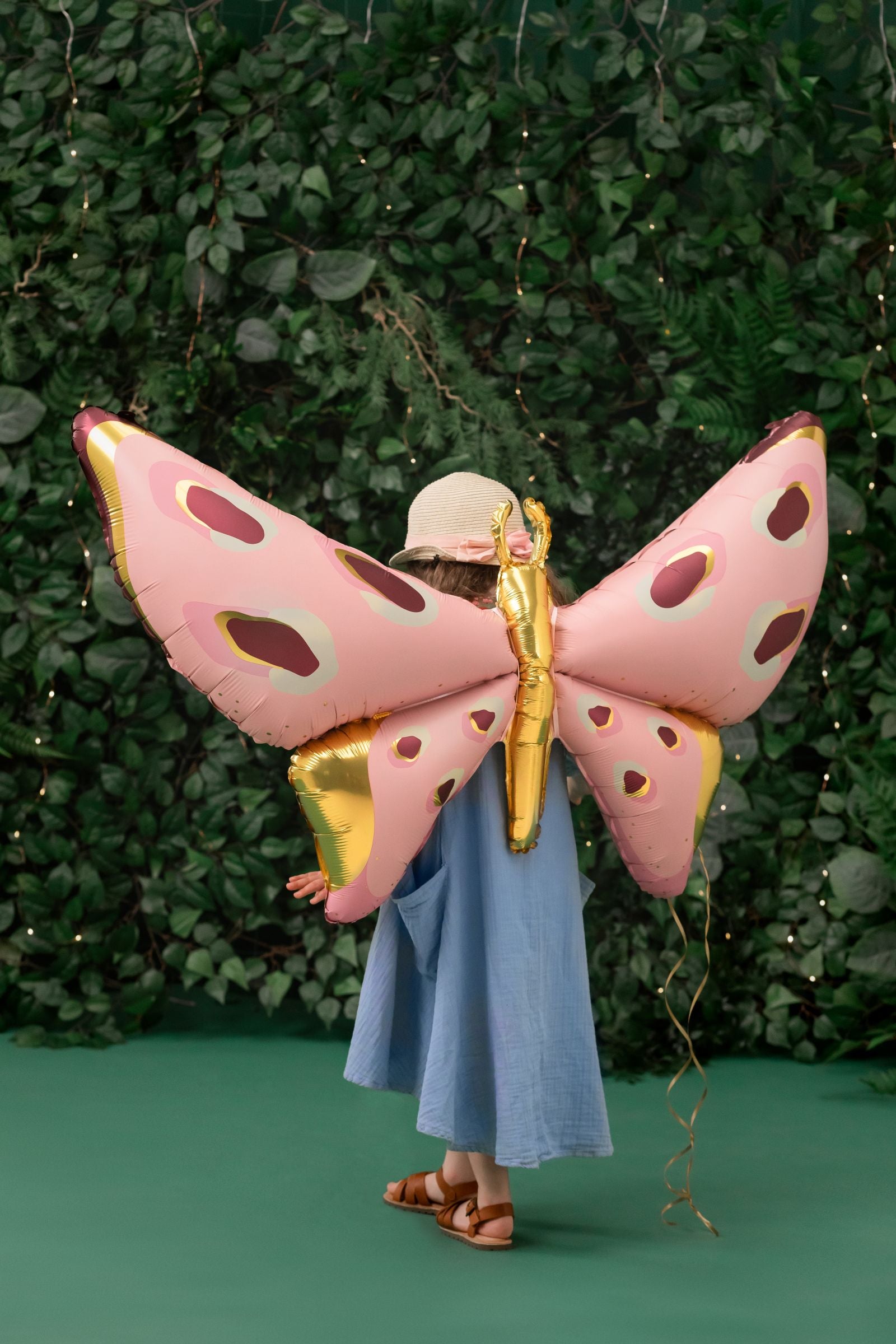 A child is turned away from the camera, holding a large pink and gold foil butterfly balloon with both hands. The balloon is attached with ribbons, appearing as wings, and sits next to a small bundle of ribbons tied at the bottom.