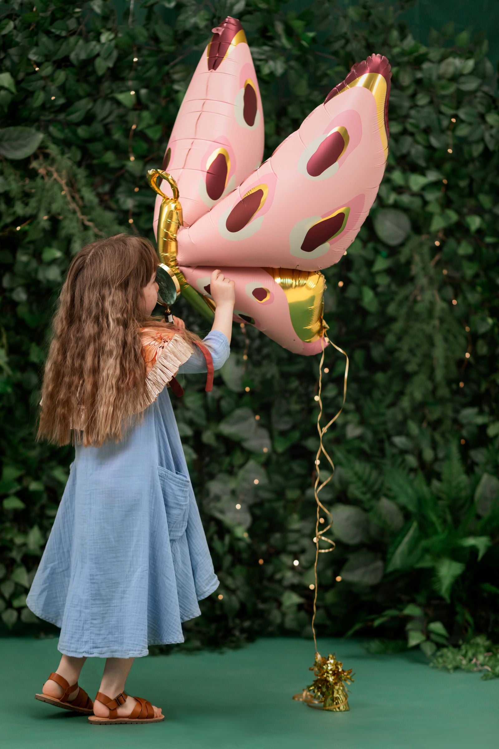 A child is standing and smiling directly at the camera, holding a large pink and gold foil butterfly balloon in one hand. The balloon is attached with ribbons, appearing like wings, with a forest-themed backdrop.