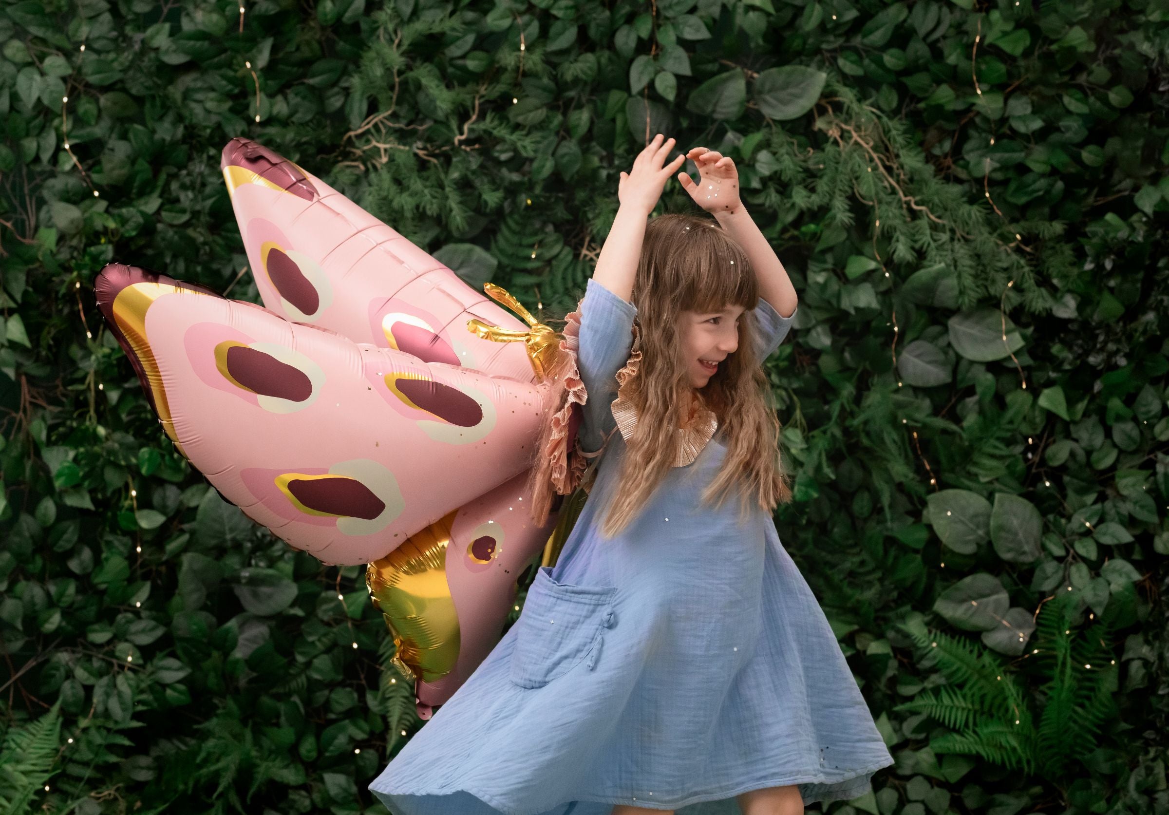 A child is playing and smiling, with a large pink, gold, and burgundy foil butterfly balloon attached to their back, as if it were a pair of wings. They are in a forest-themed room with a lush green backdrop.
