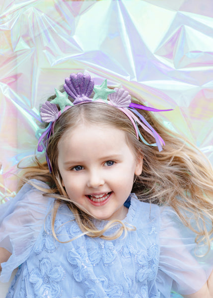 A smiling child with long light brown hair wearing a glittery mermaid-themed headband adorned with purple seashells, mint green starfish, and multi-colored ribbons, set against an iridescent, shimmery light purple and pink background.