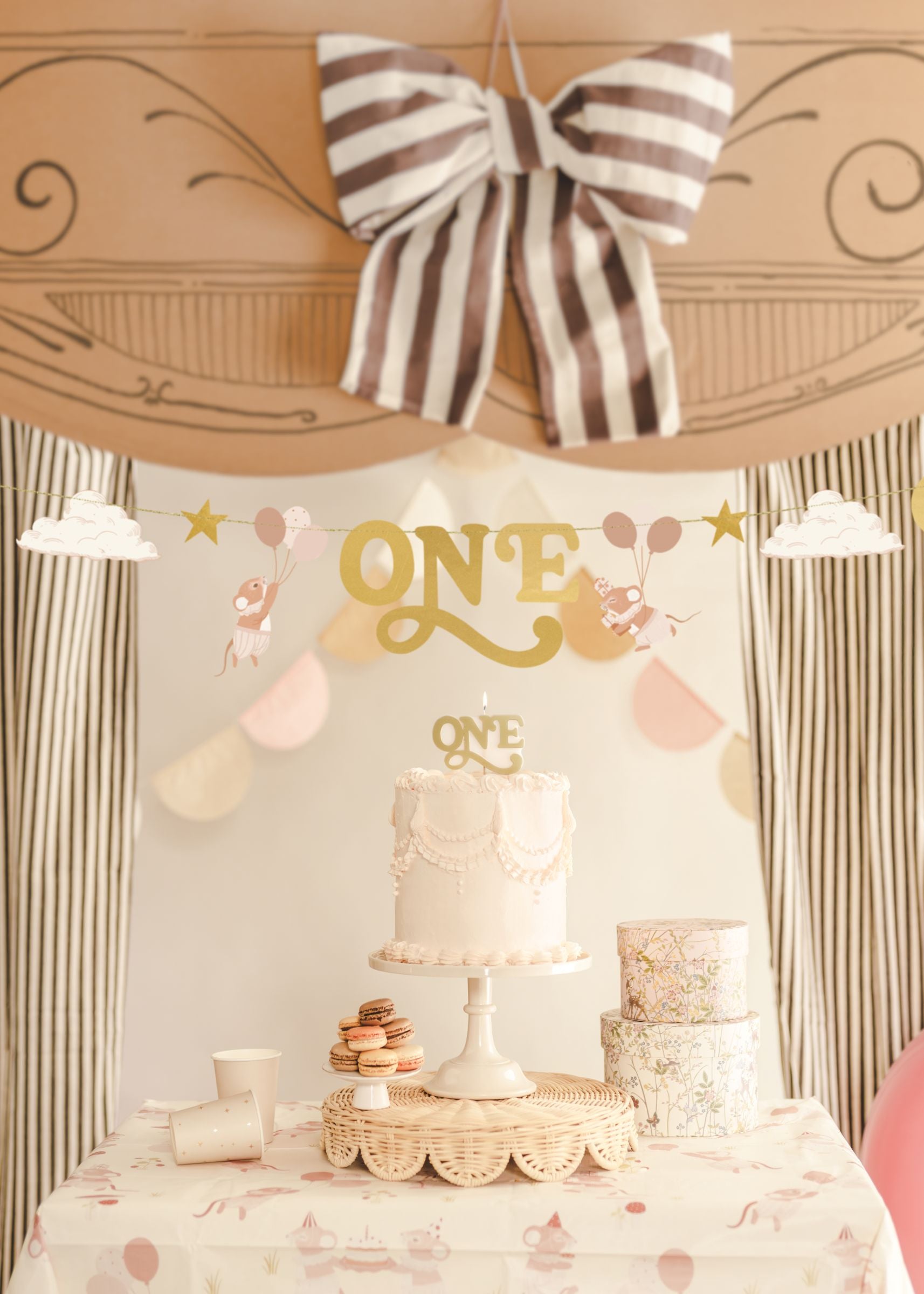 A beautifully styled 1st birthday dessert table featuring a white cake, macarons, and a beige mice-themed 'ONE' banner with gold stars hung gracefully above.
