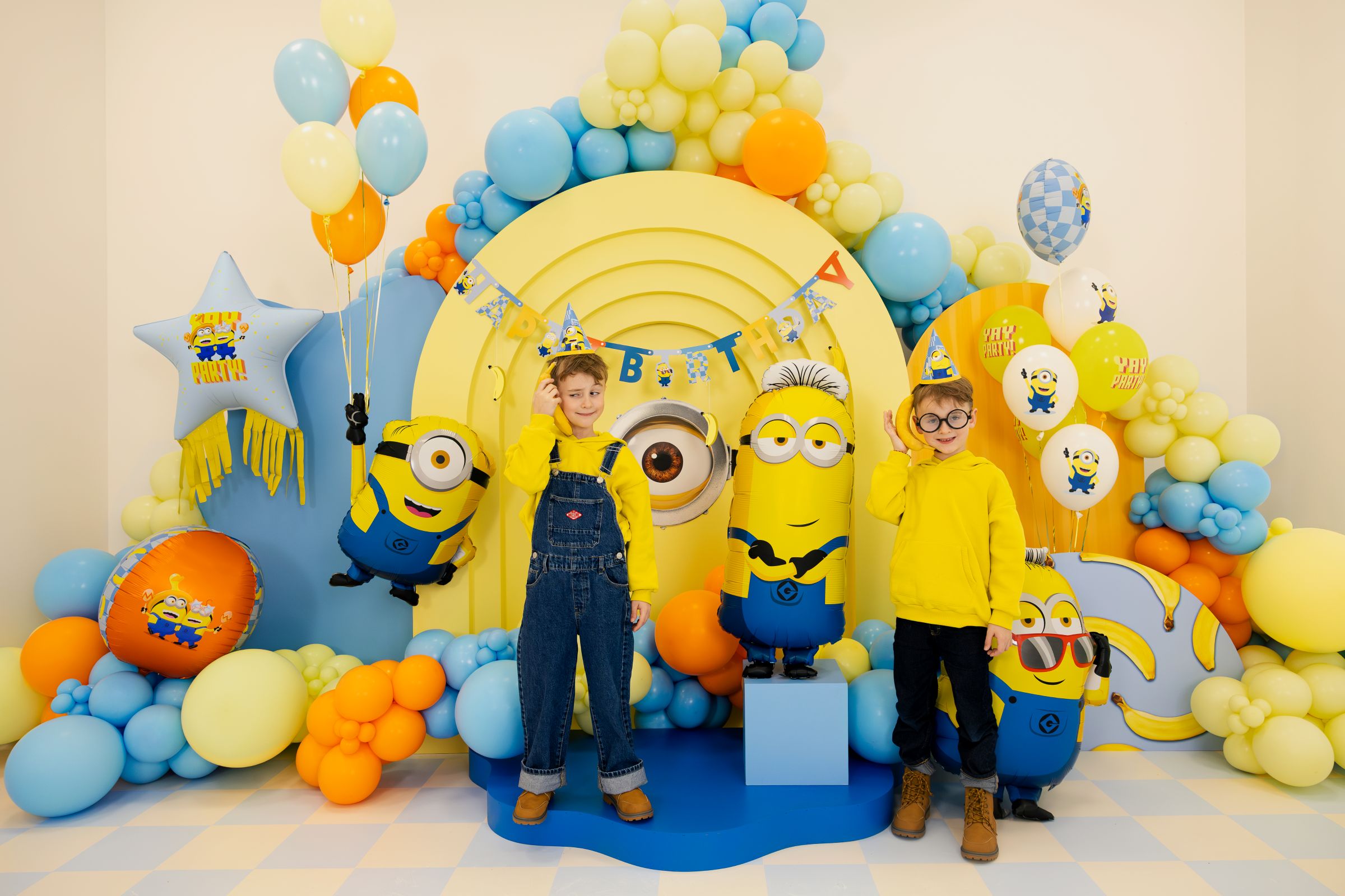 Two children in front of a colorful Minions-themed birthday backdrop with balloons and Minion Mel floating.