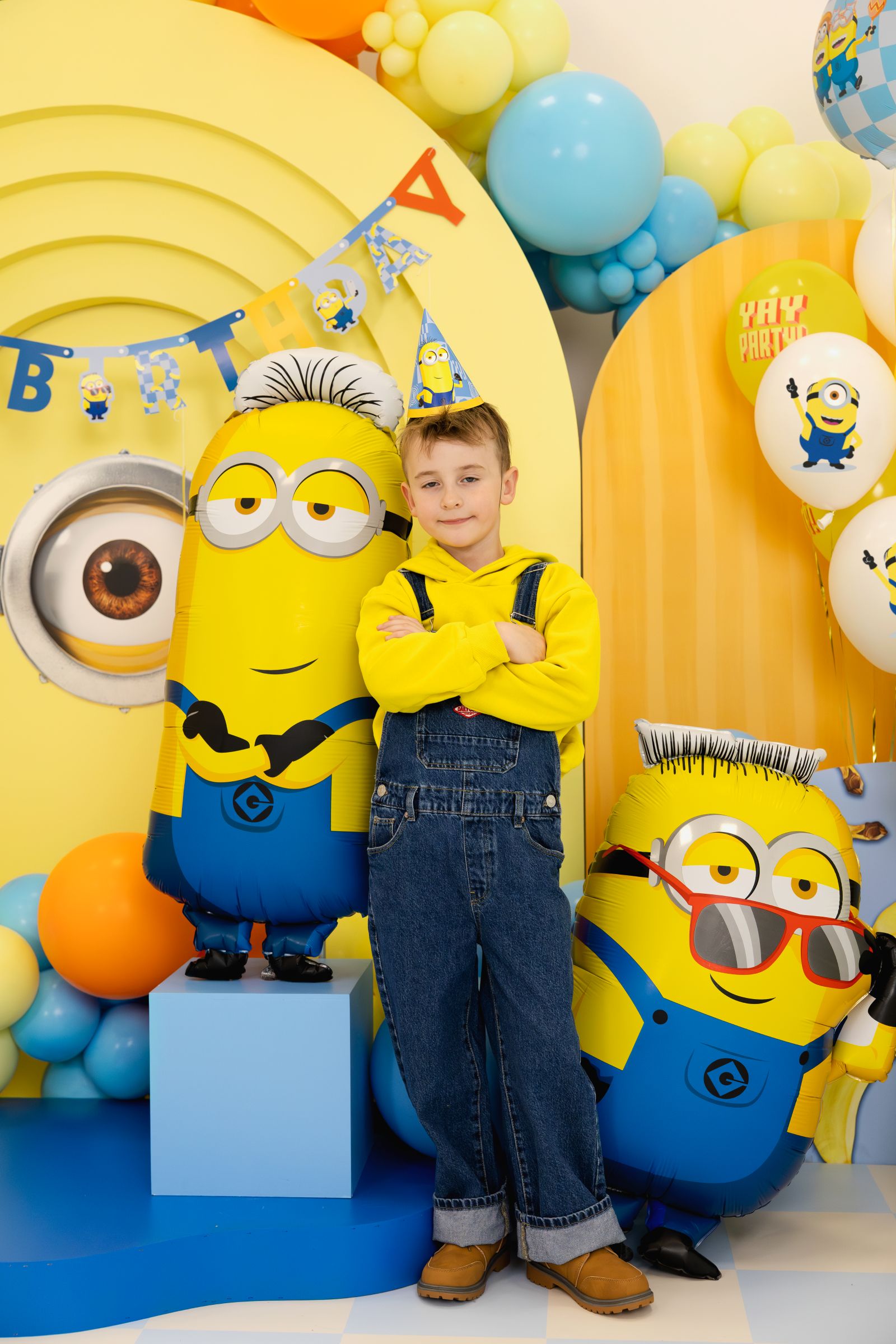 A child in denim overalls posing next to a giant Minion Tim foil balloon at a fully decorated birthday party.