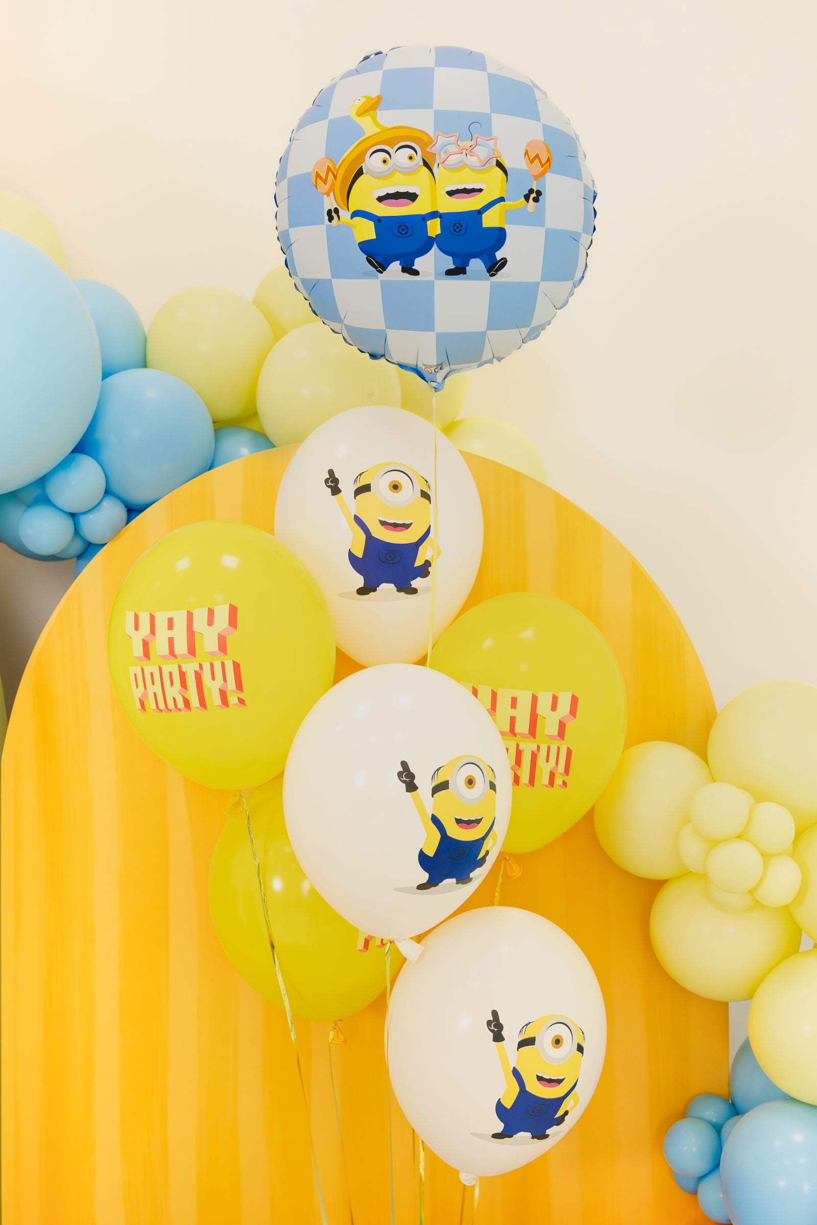 A party setup featuring a blue and white checkered Minions foil balloon floating above a cluster of yellow and white latex balloons, some printed with Minions and 'YAY PARTY!'
