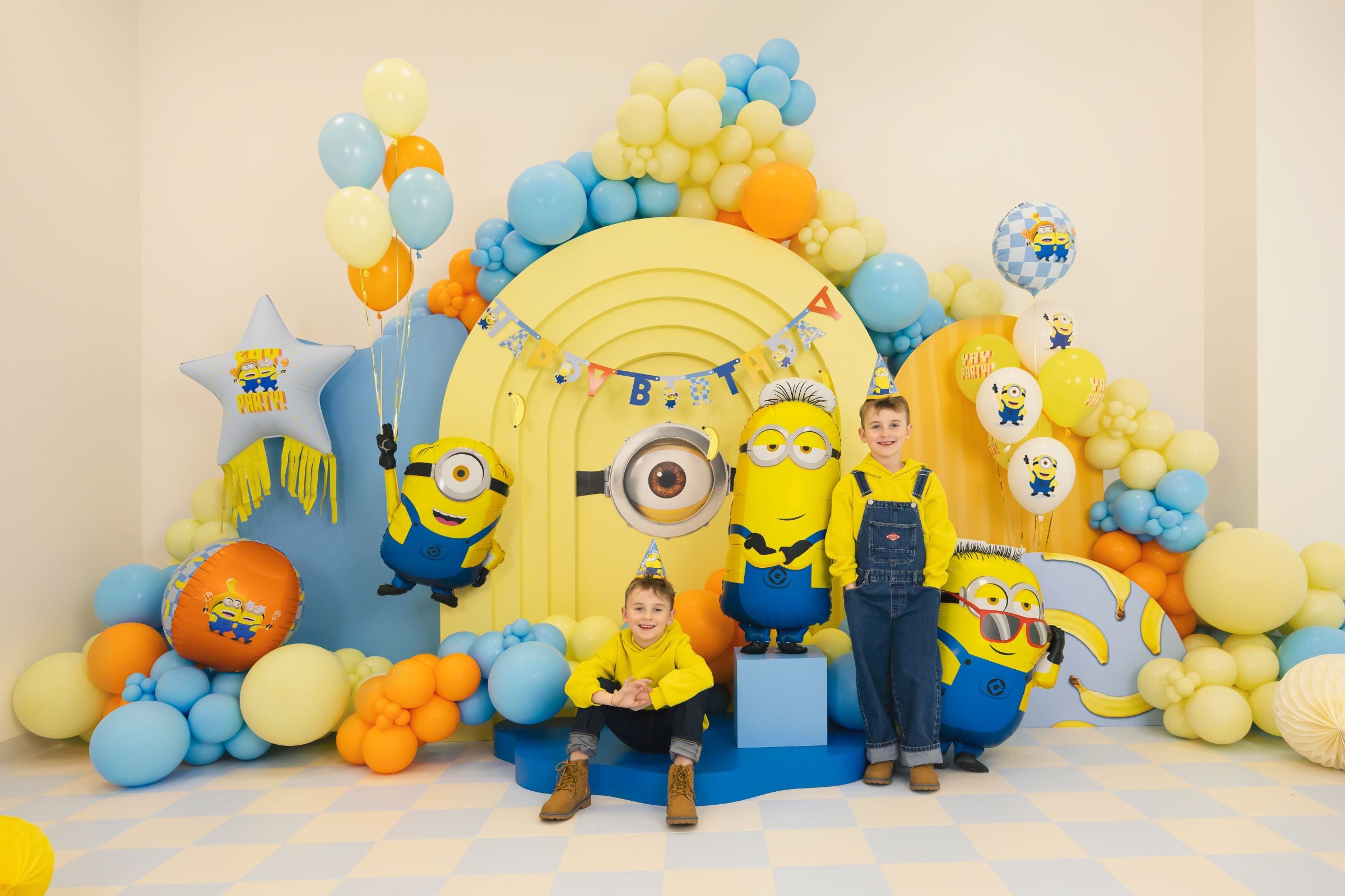 Two children in Minions outfits posing in front of a massive Minions-themed party backdrop featuring large foil Minion balloons, balloon arches, and a giant Minion eye.