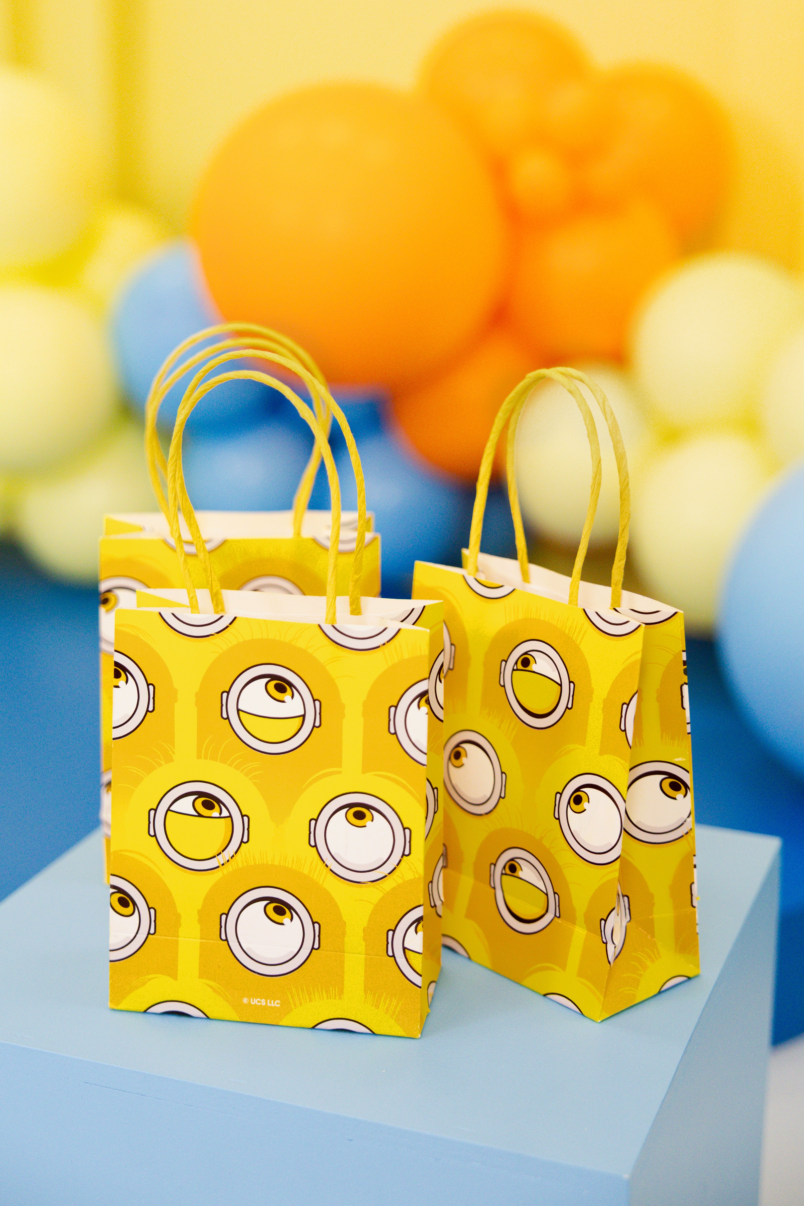 Three yellow Minions paper gift bags sitting on a light blue display block, with a vibrant arch of colorful party balloons slightly out of focus in the background.
