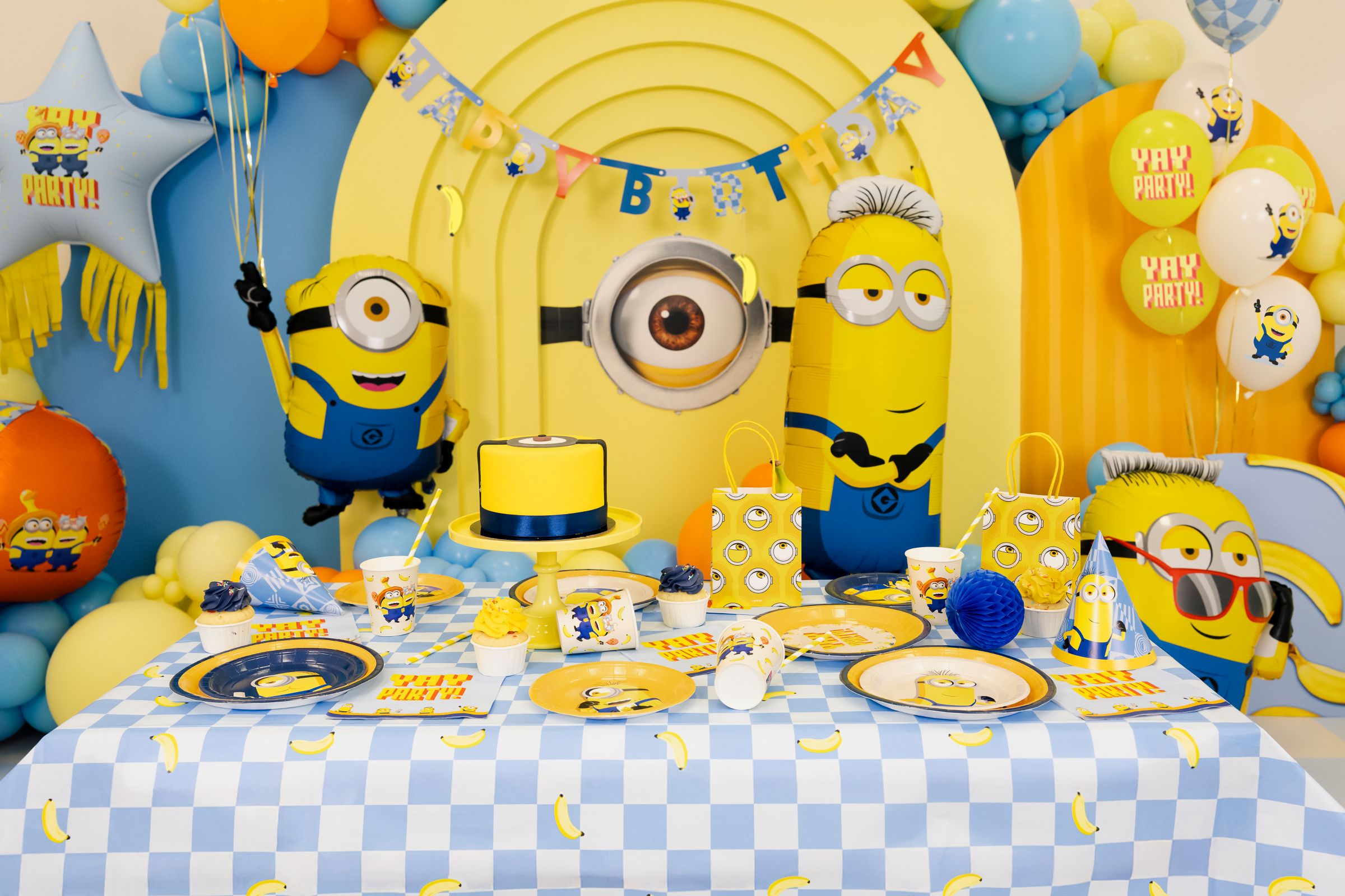 A fully decorated Minions-themed party table featuring a checkered tablecloth, matching paper plates, cups, napkins, a yellow cake, and Minions paper gift bags, set against a fun backdrop of colorful balloons and giant Minion character foil balloons.