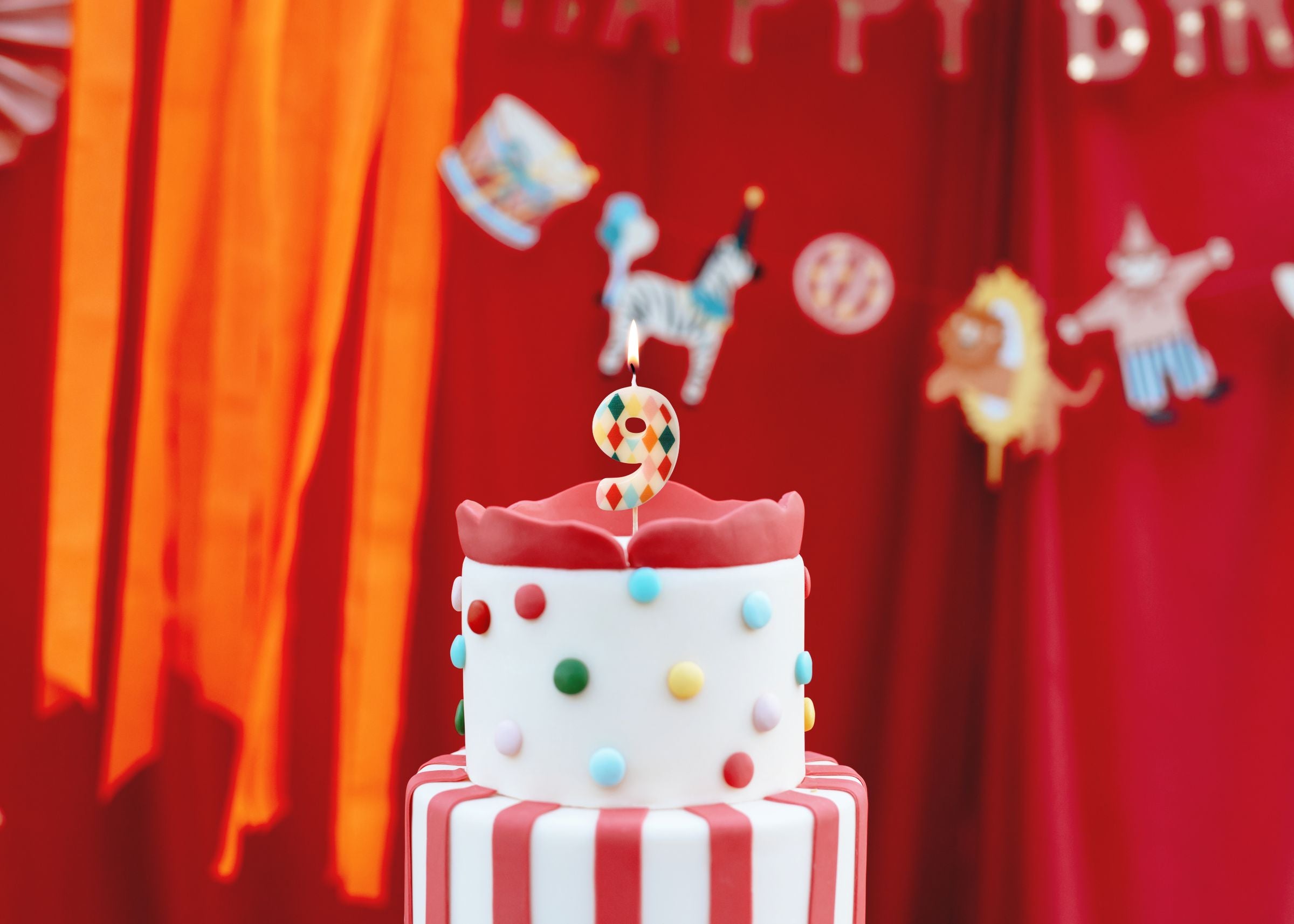 A lit number 9 rhombus patterned candle sitting on top of a red and white striped circus-themed cake.