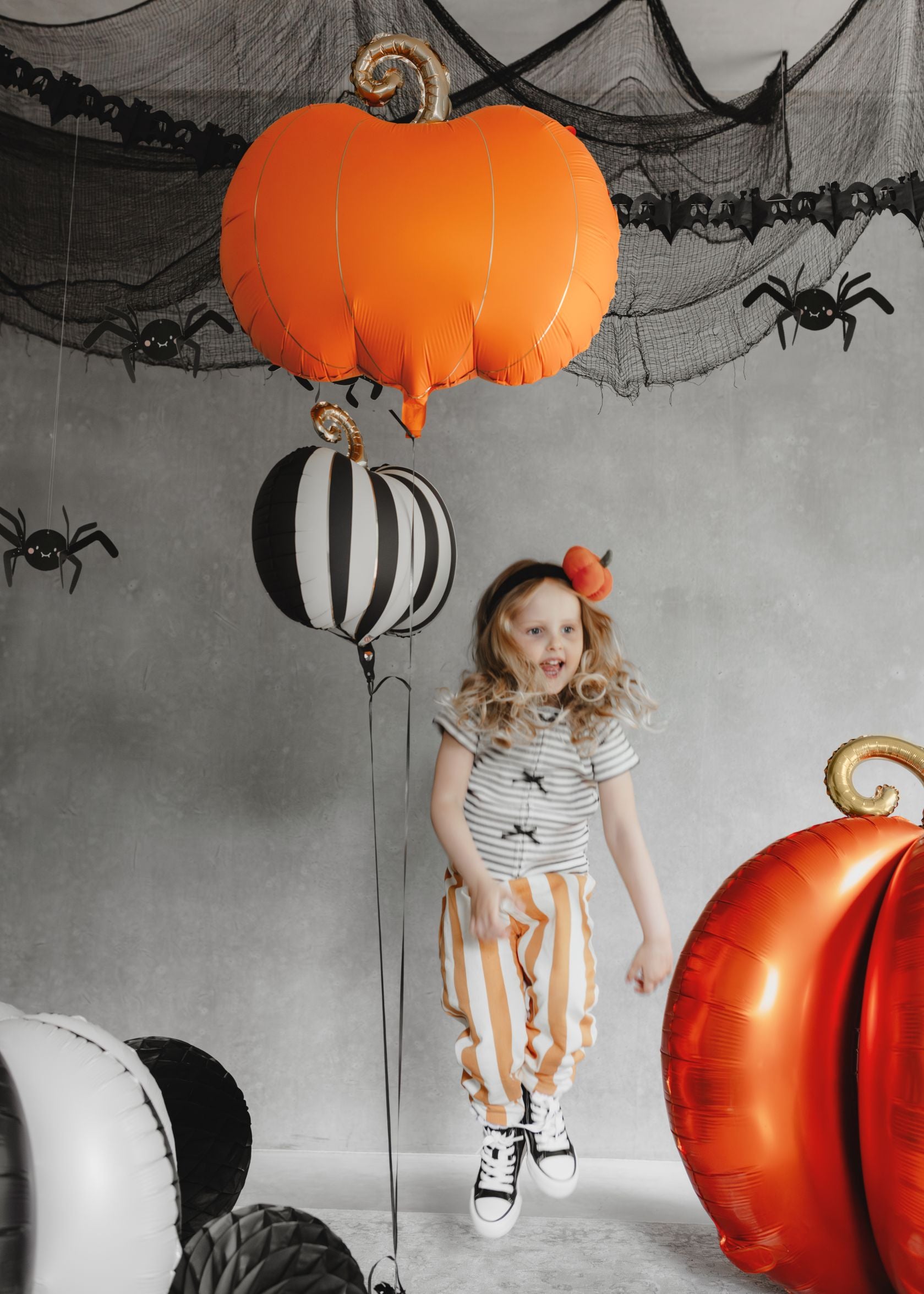 A child jumping in a Halloween-decorated room. Large orange and gold pumpkin foil balloons are visible, along with a black and white striped pumpkin balloon. The background features a grey wall with cobweb netting and spider decorations.