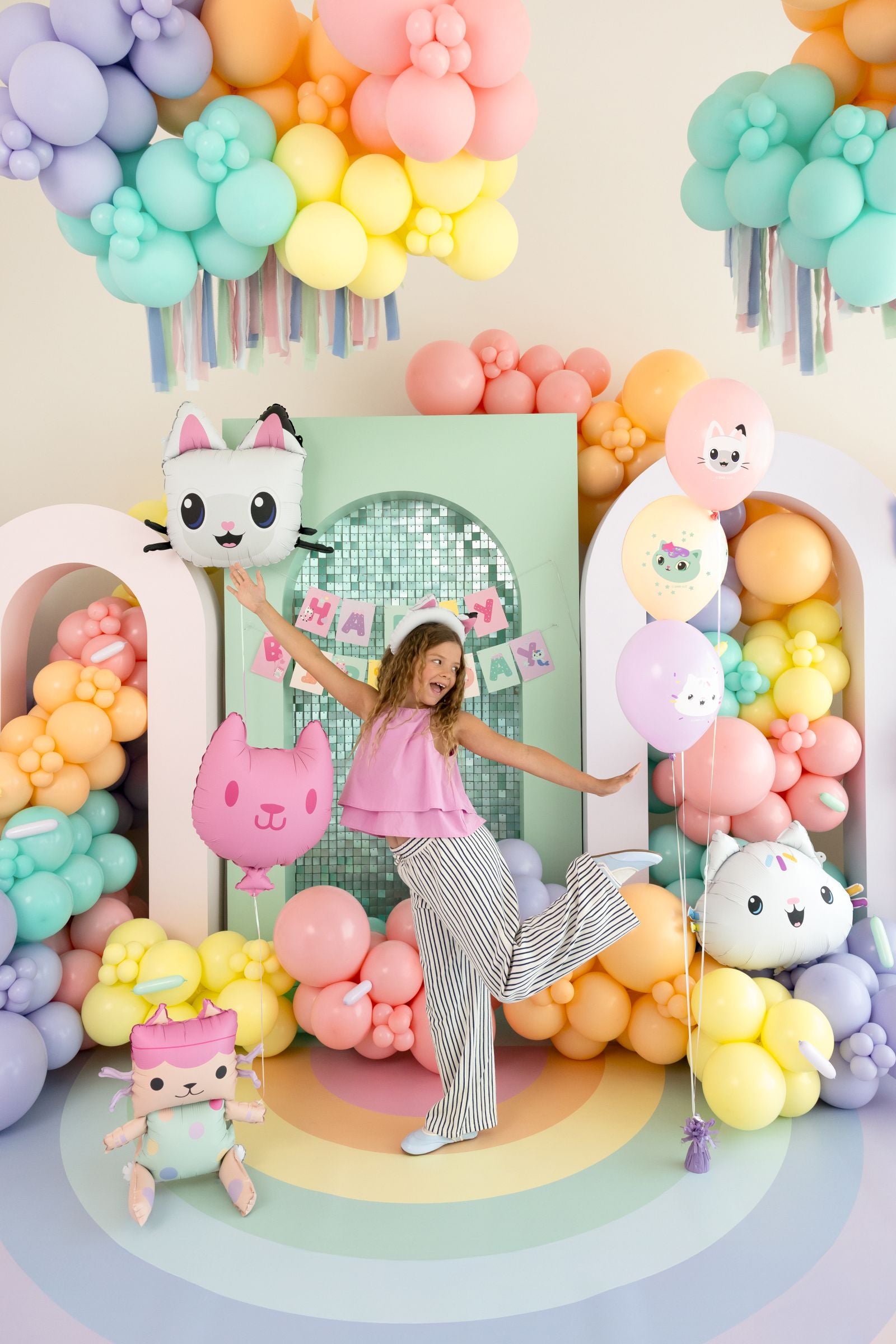 Child posing in front of a Gabby's Dollhouse birthday party scene including colorful balloon arch with cartoon cat figures.