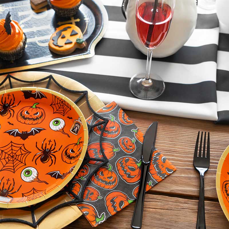 A Halloween party table setting with an orange spiderweb plate, black cutlery, and a black paper napkin printed with scary orange pumpkins.