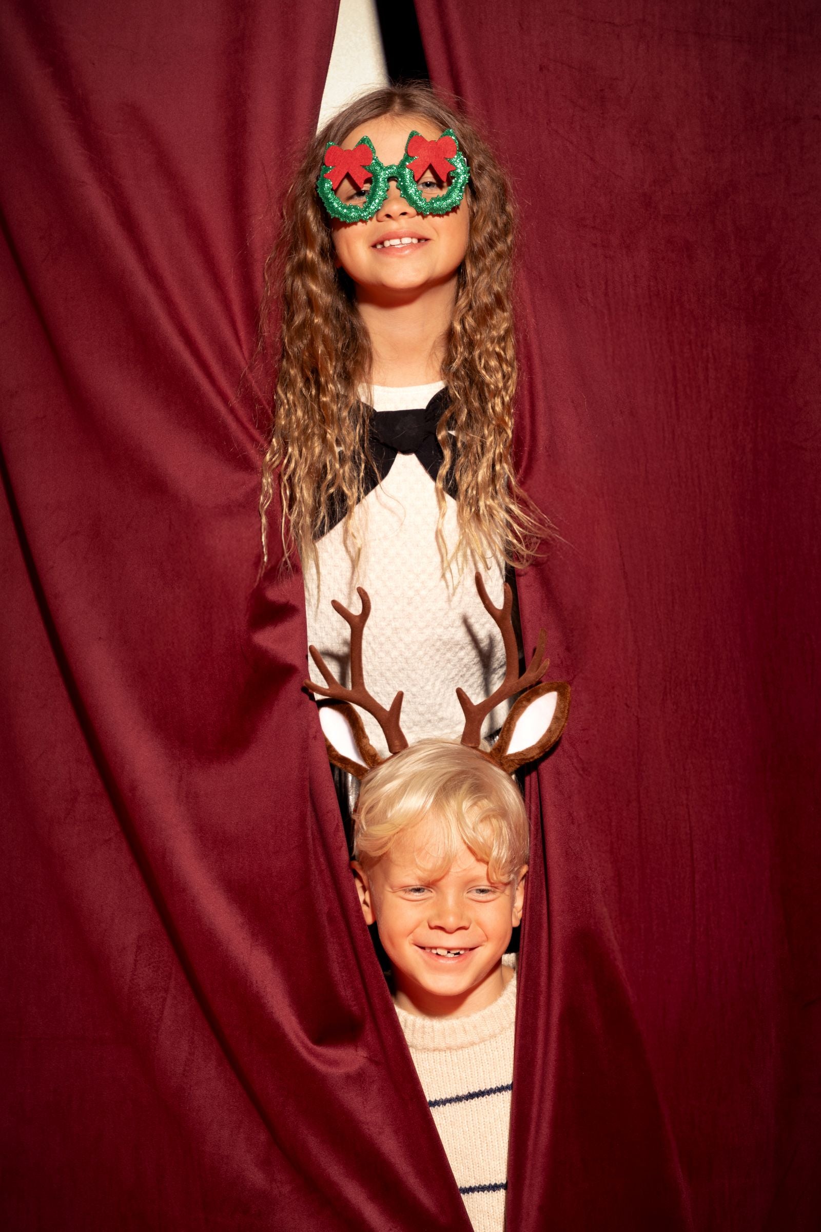 Two children peeking through a red curtain; the child on the bottom is wearing the reindeer antler headband.