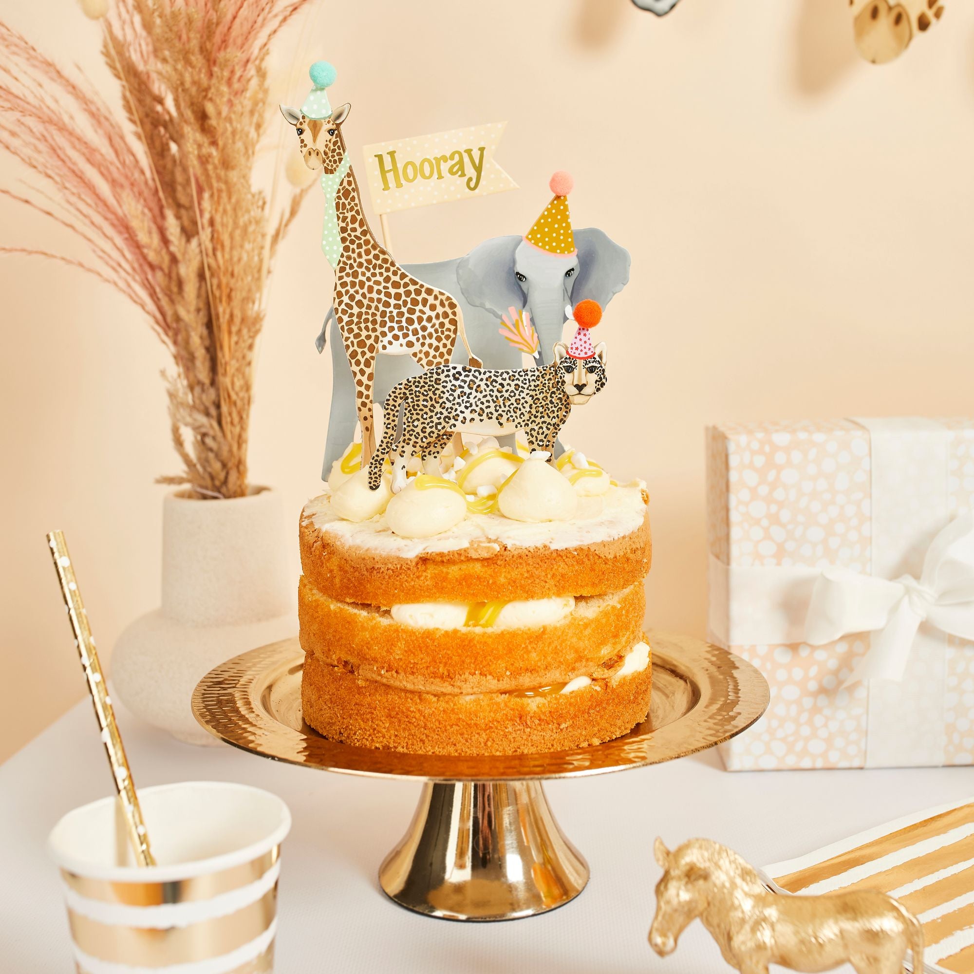 A birthday cake topped with safari animal toppers including an elephant, giraffe, and leopard wearing 3D pom-pom hats.