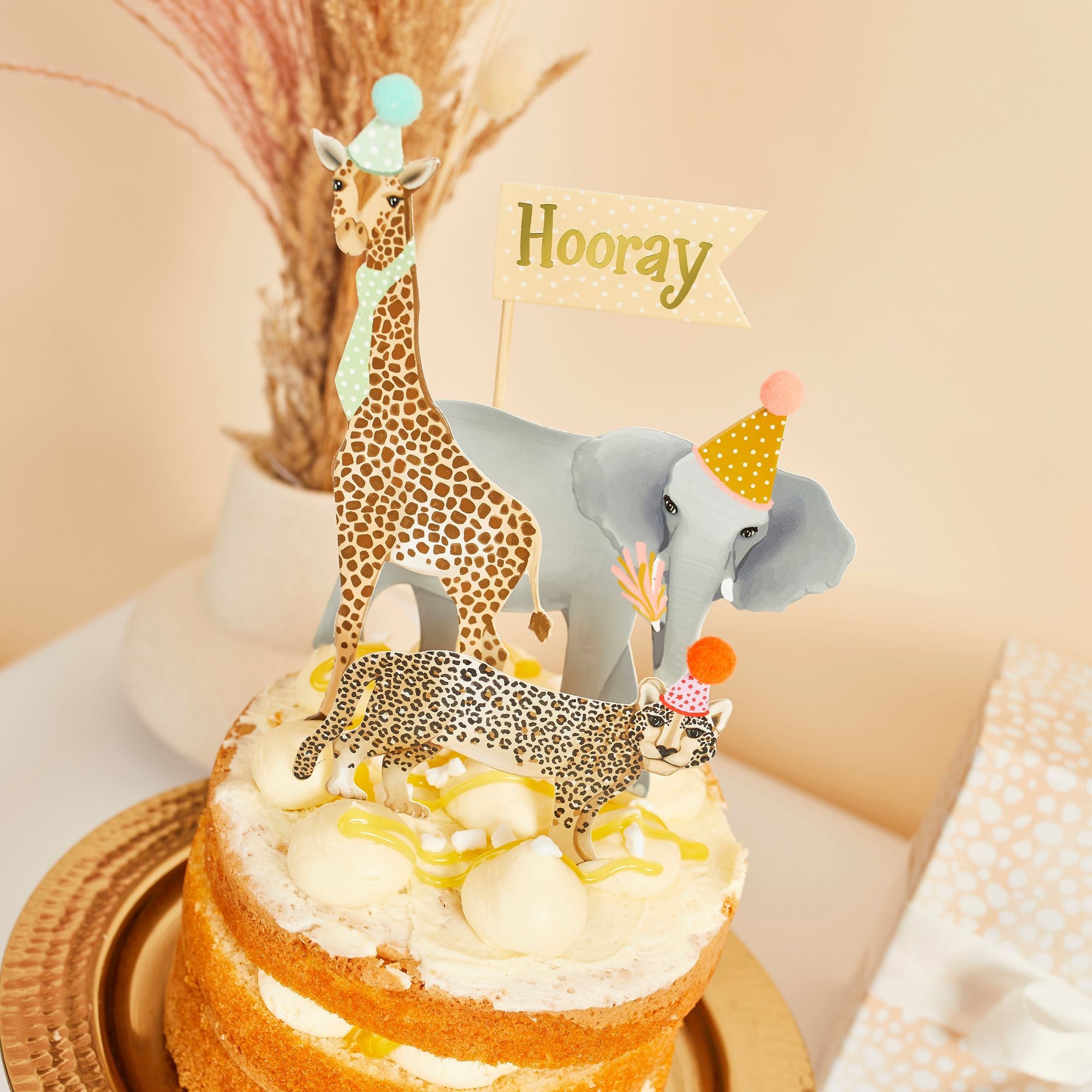 Close-up of a naked tiered cake with lemon curd, decorated with safari animal food picks featuring detailed illustrations and pom-pom hats.