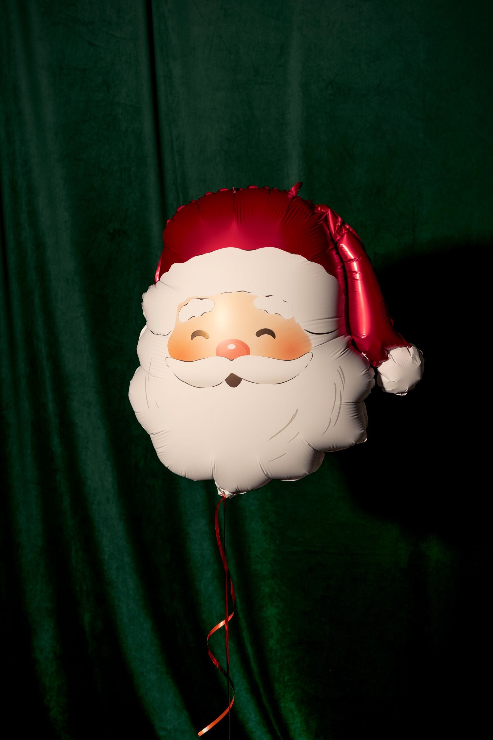 A large Santa Claus head foil balloon floating against a dark green velvet curtain.