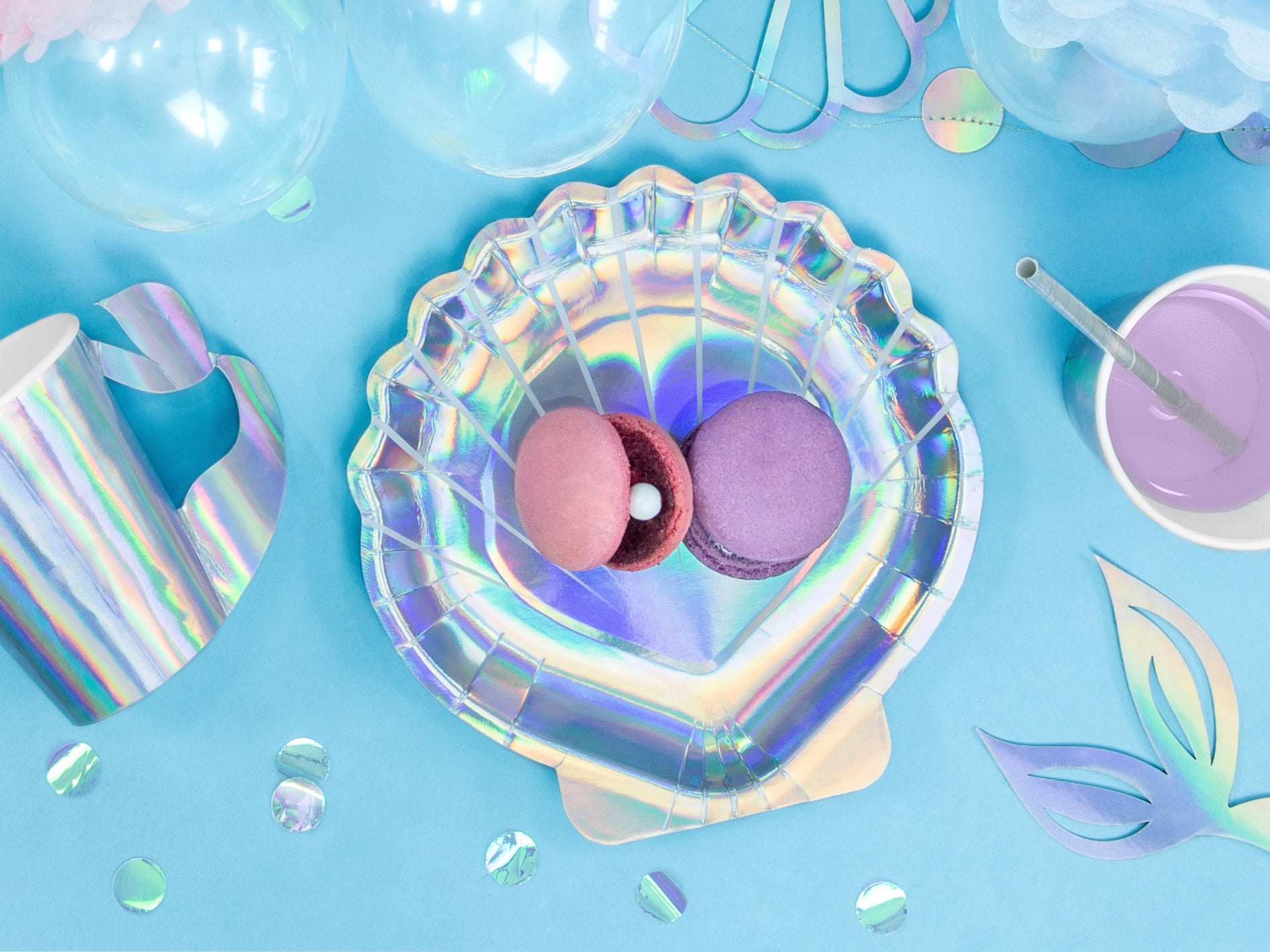 An overhead view of an iridescent seashell-shaped paper plate holding a pink and a purple macaron with a pearl, surrounded by iridescent mermaid-themed party supplies like a matching cup, clear balloons, and confetti, all on a light blue background.
