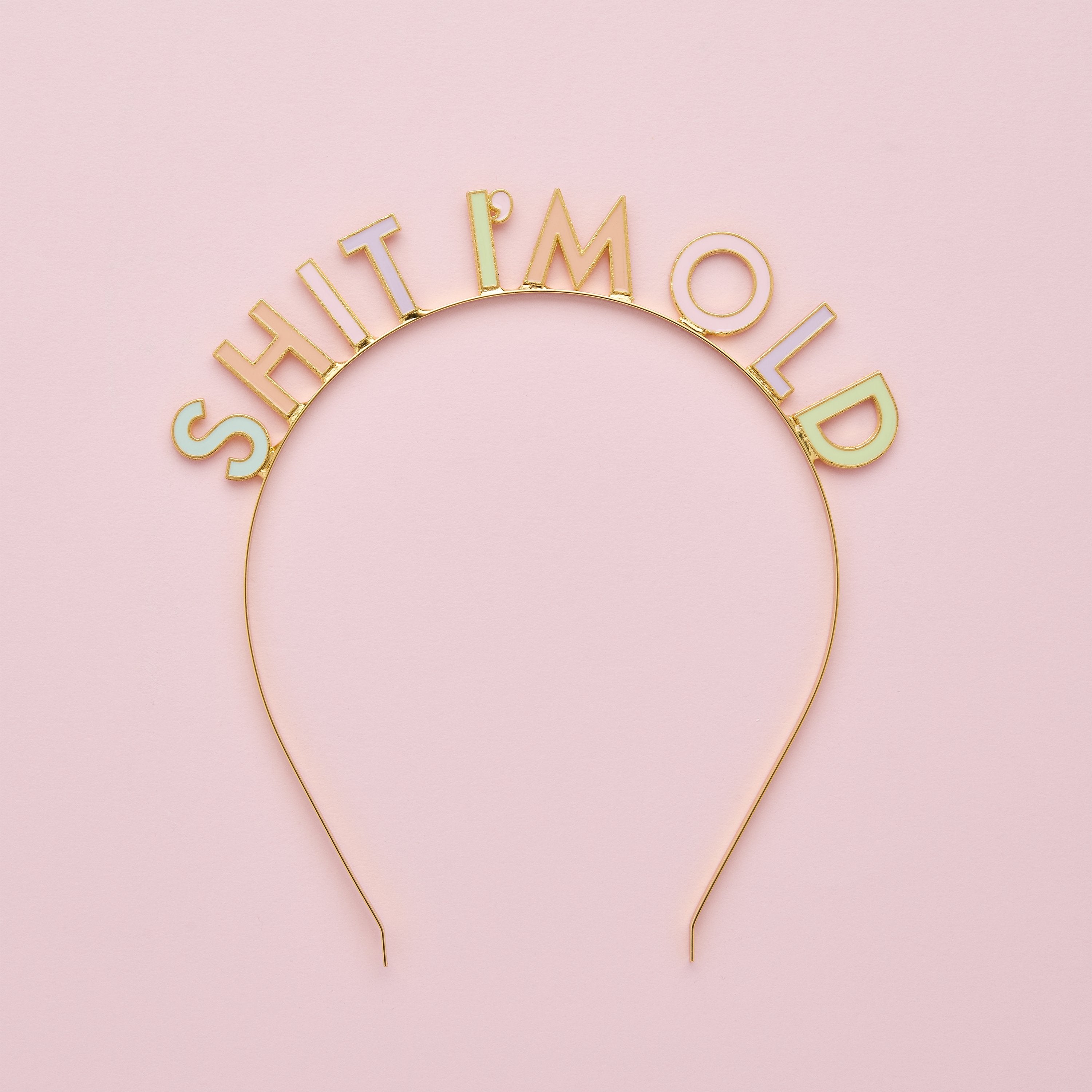 A gold novelty birthday tiara reading "SHIT I'M OLD" with pastel enamel lettering, displayed flat against a clean, soft pink background.