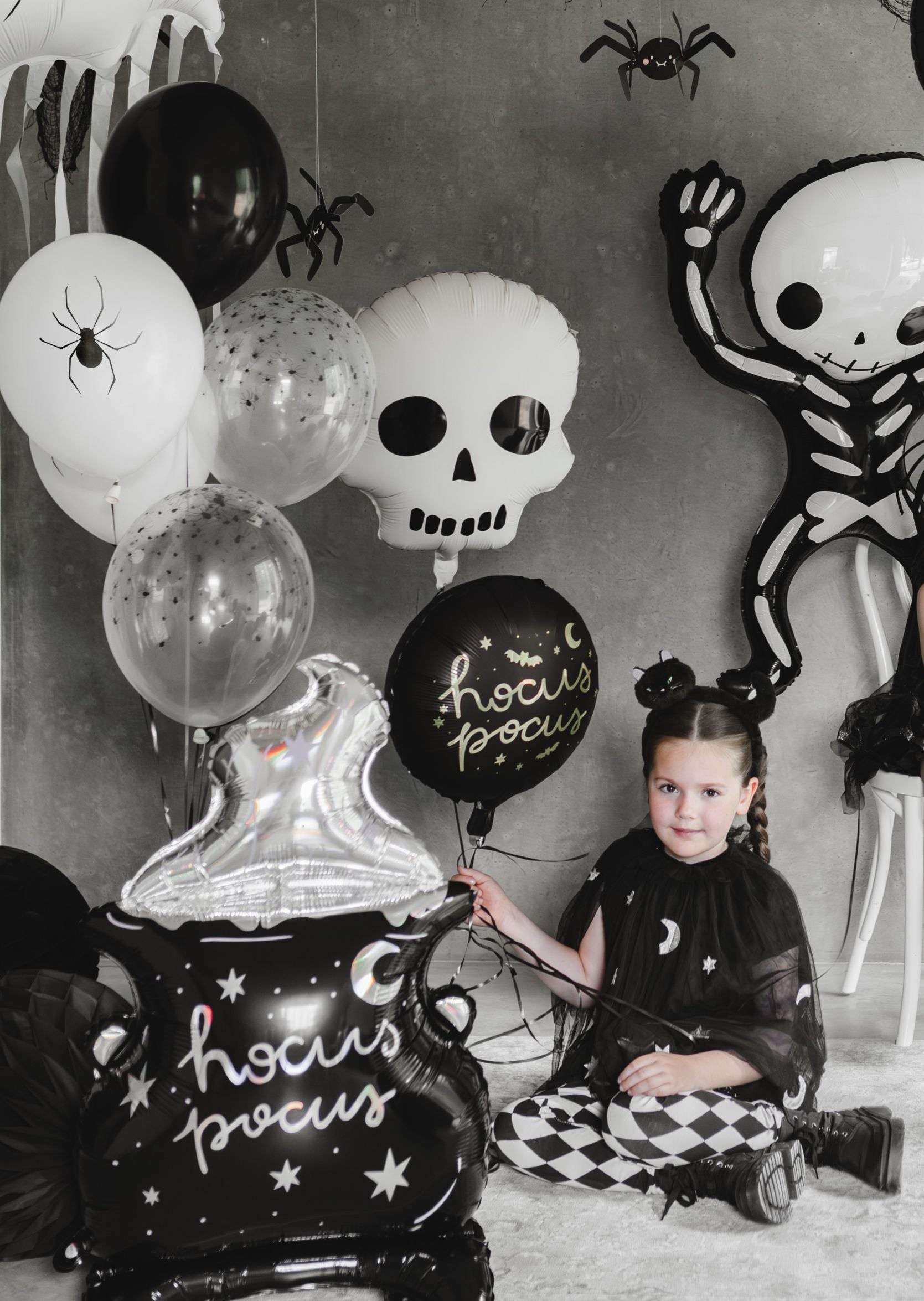 A Halloween party setup with a child in a black cape and checkered pants sitting on the floor amidst black and white balloons. Decorations include spider balloons, a white skull foil balloon, a skeleton foil balloon, and black cauldron-shaped balloons that say "hocus pocus."