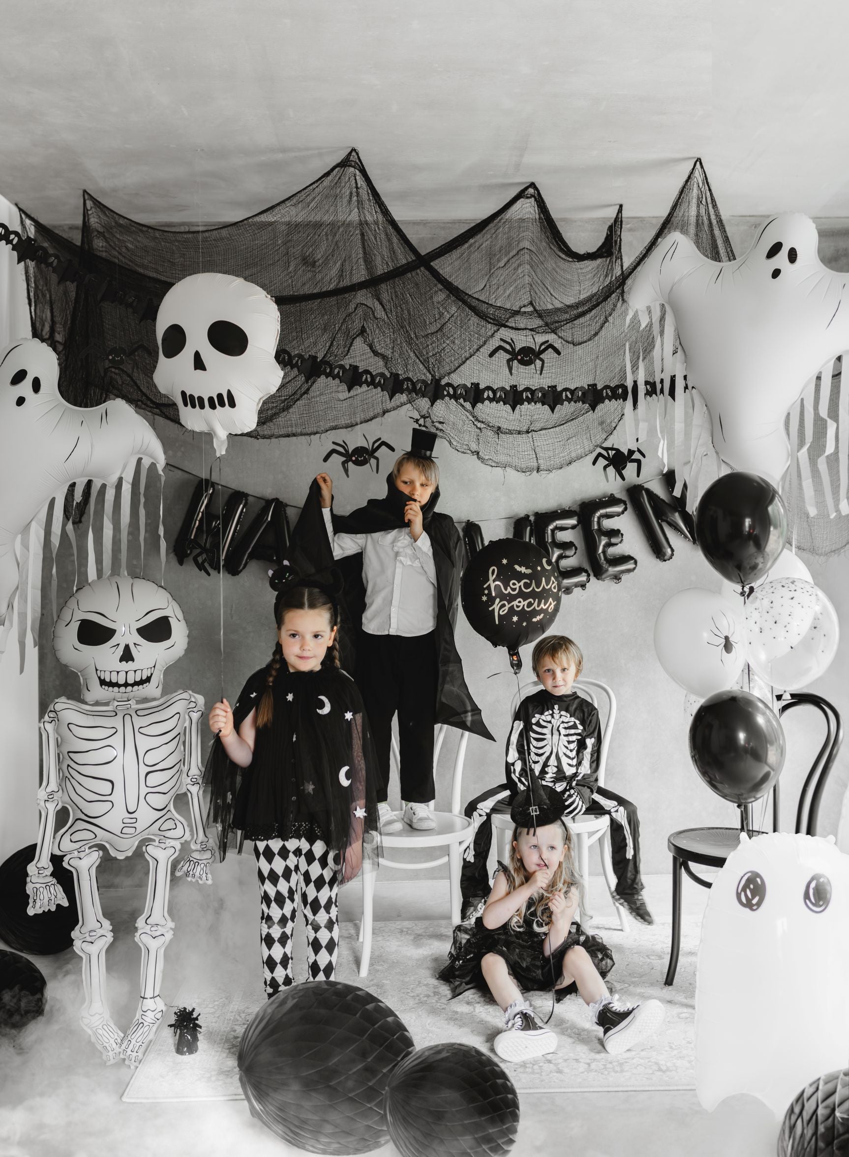 A Halloween party scene with several children in costumes (skeleton, witch, vampire) posing around black and white decorations. Large white skull and ghost foil balloons, a skeleton foil balloon, black netting, and honeycomb balls create a spooky atmosphere.