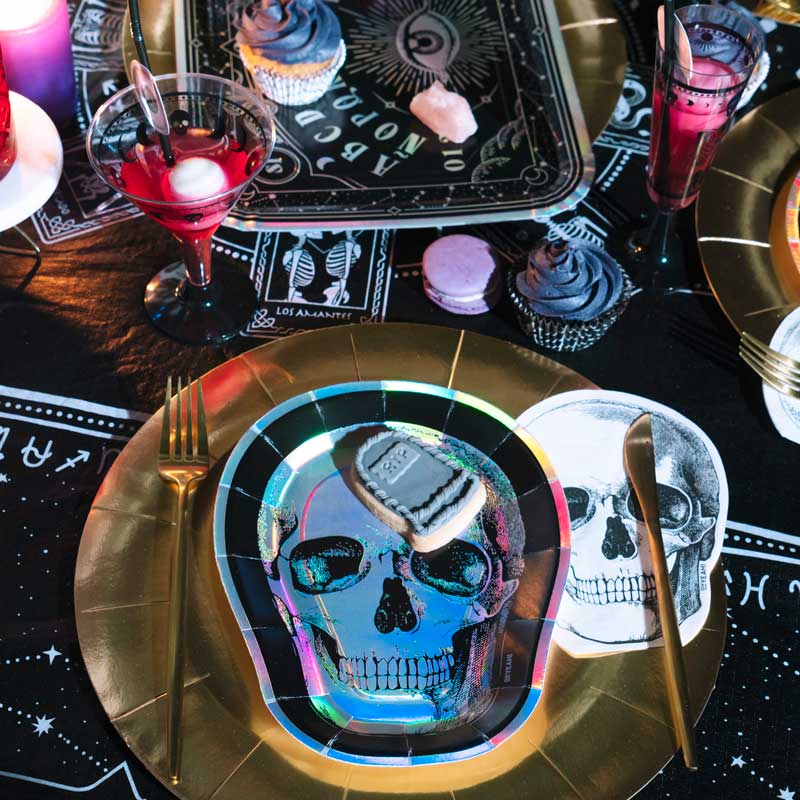 A dark Halloween party table setting with an iridescent skull plate, black cutlery, a cocktail, and a white skull-shaped paper napkin.