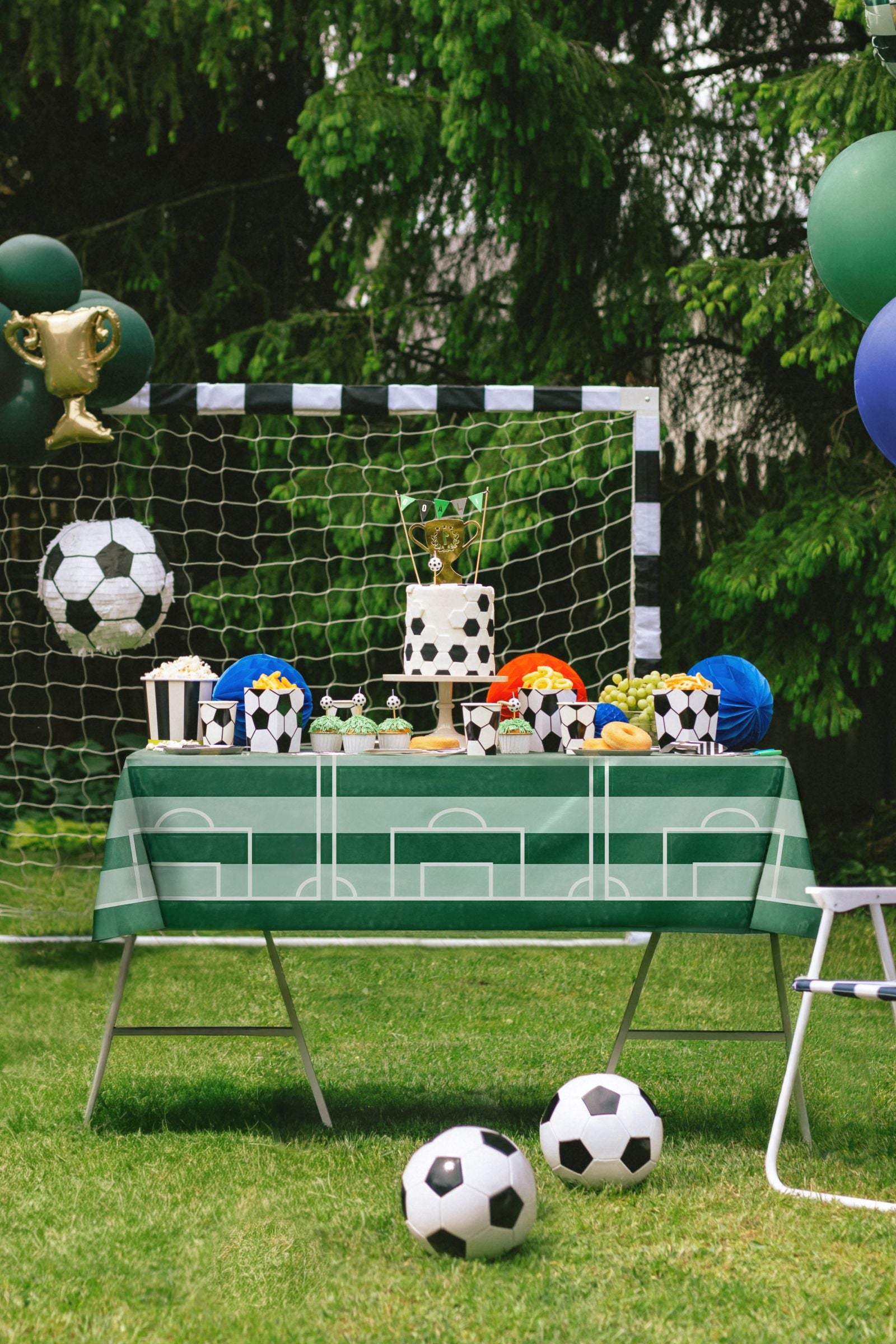 Children's party setup with soccer-themed decorations, including a table with soccer balls and trophies, and a goal in the background.
