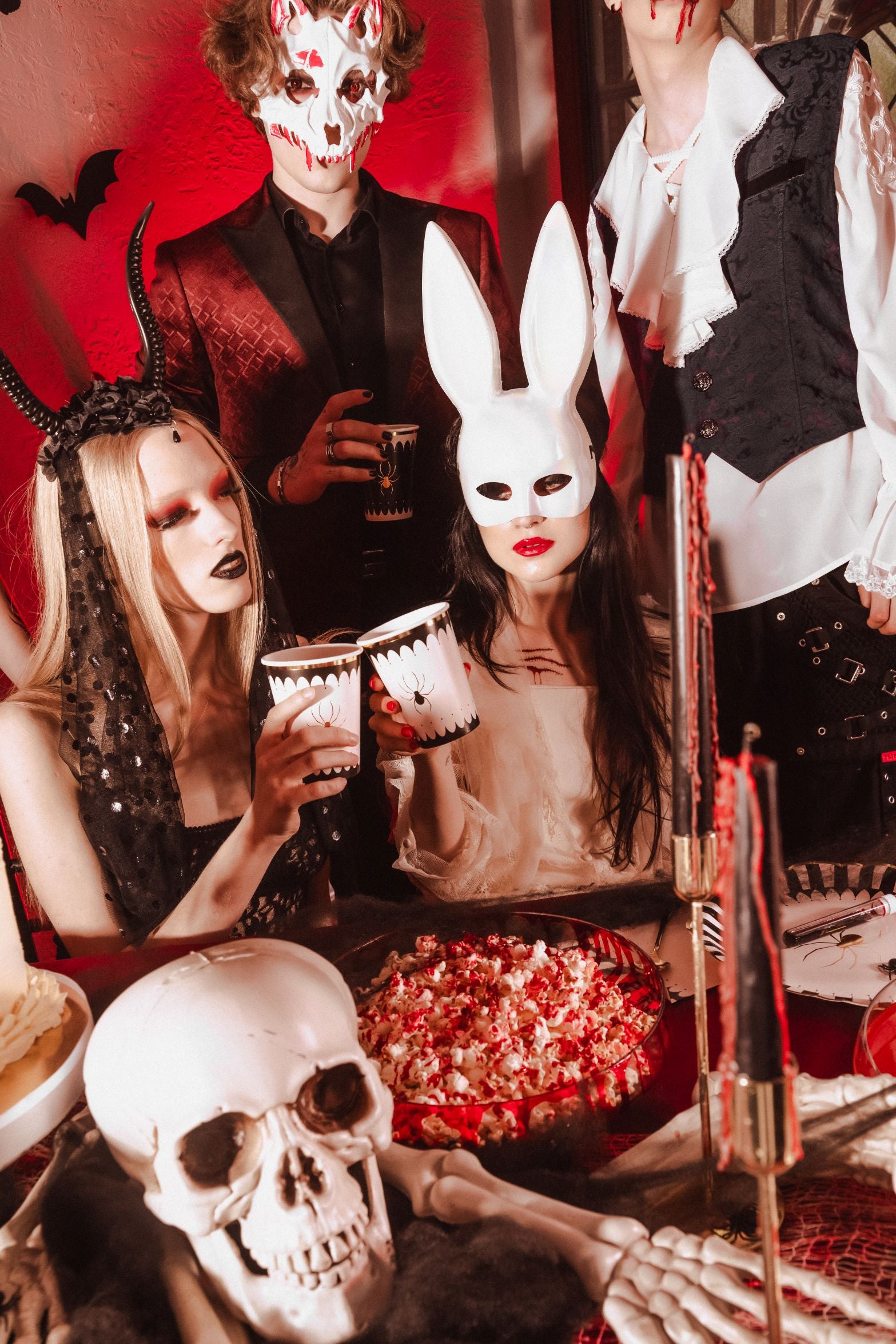 A group of people in various Halloween costumes and masks (including a skull mask and bunny mask) at a party table, holding up spider-themed cups. The table is decorated with a skull, popcorn in a bowl, and black candles.