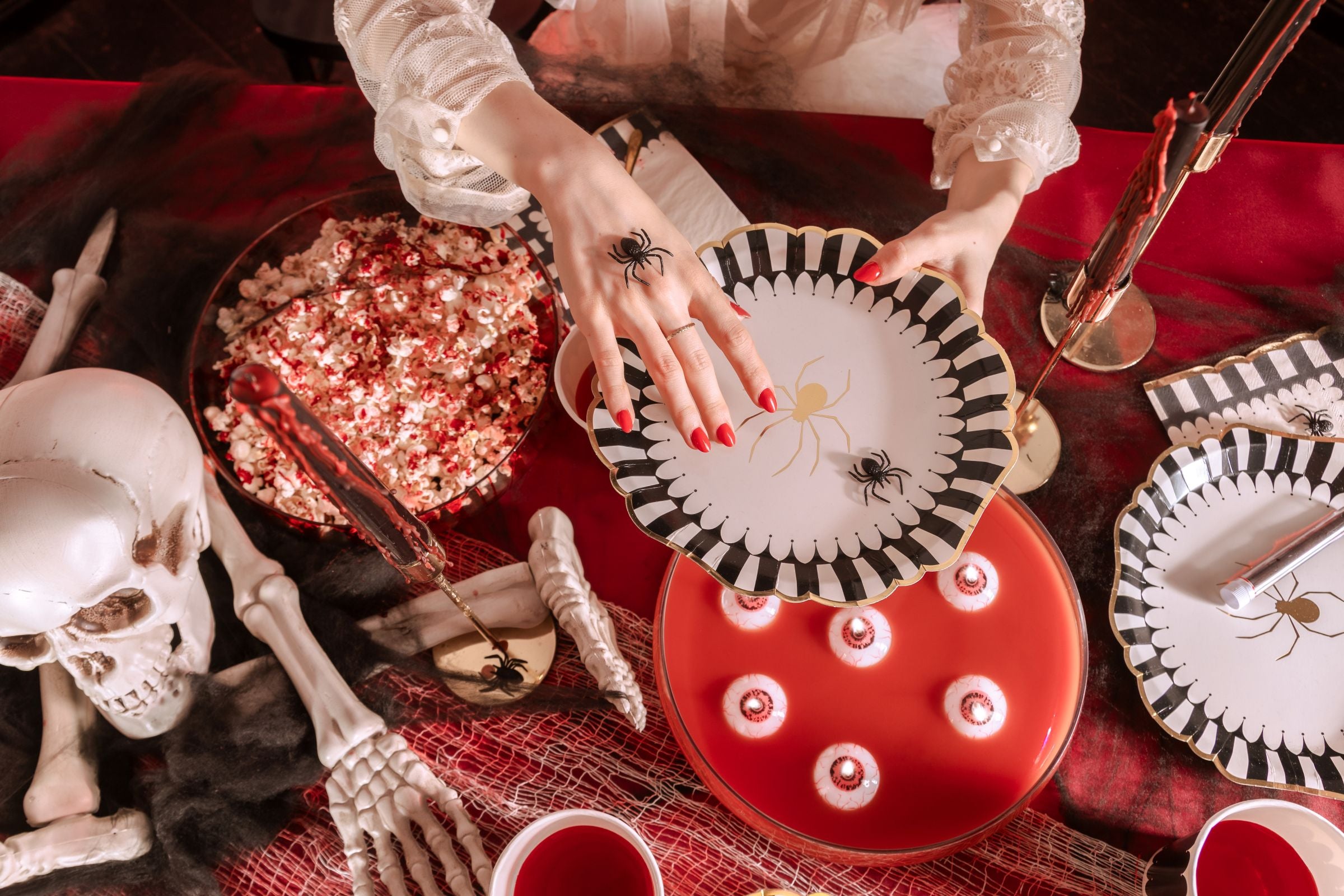 An overhead view of a Halloween-themed table decorated with a red tablecloth and black netting. Hands with red nail polish are placing or holding a white plate with a black and white scalloped edge and a gold spider design, above a red punch bowl with floating "eyeball" candles. Other decorations include a skeleton skull and bones, popcorn, and themed cups.