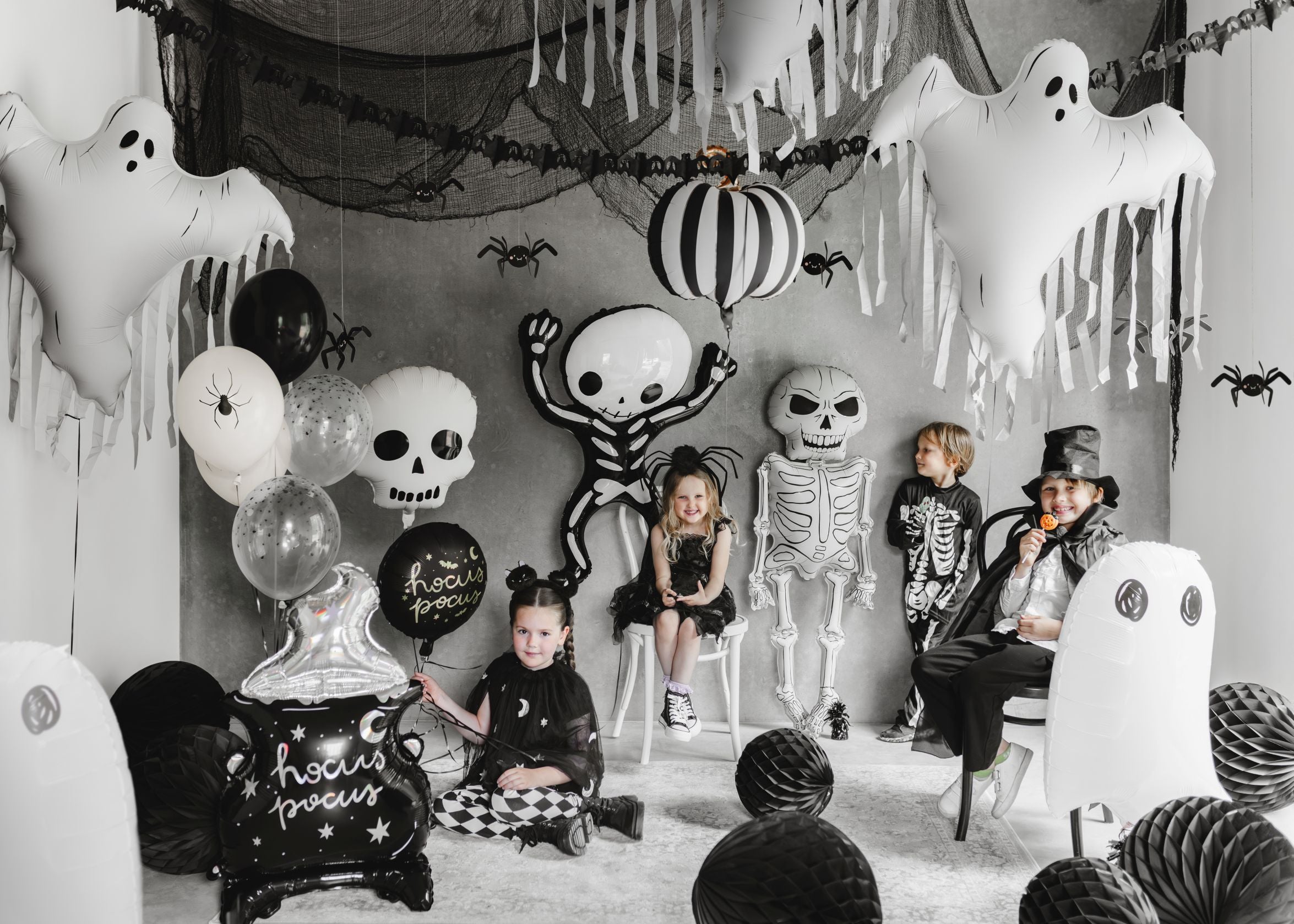 A Halloween party scene with several children in various costumes (skeleton, harlequin, witch, wizard) posing amidst abundant decorations. The room features black netting, ghost balloons, skeleton balloons, a black and white striped pumpkin balloon, and clusters of black, white, and spider-themed balloons.