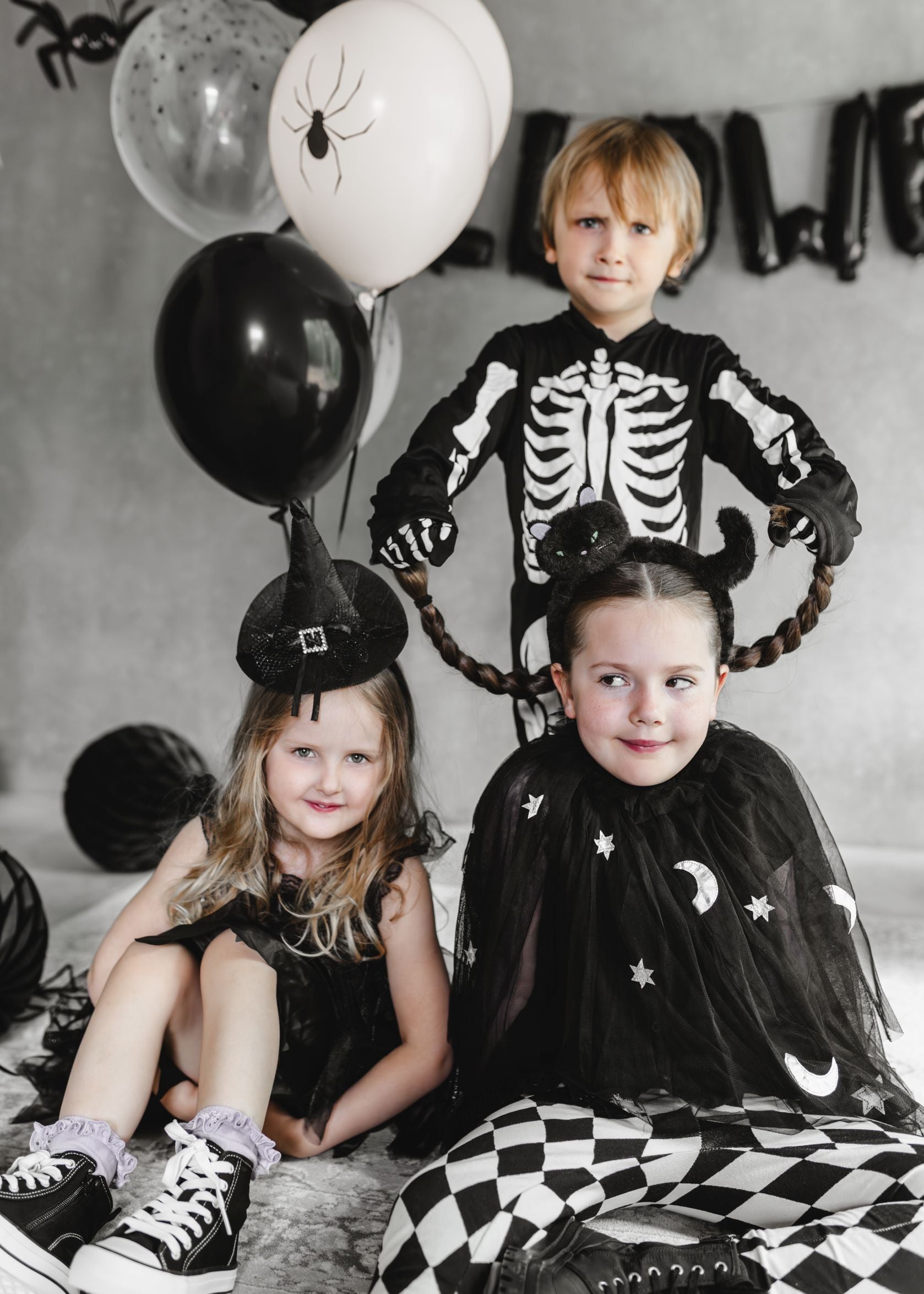 Three children in Halloween costumes (skeleton, witch, vampire-style) posing in a decorated room with black and white balloons, including spider-themed ones, black cobweb netting, and honeycomb balls.