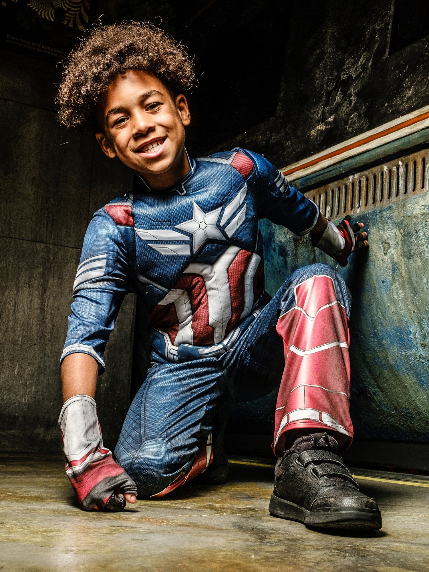 A child in a Captain America costume, without the mask, kneeling and smiling heroically, in front of a grungy, metallic background.