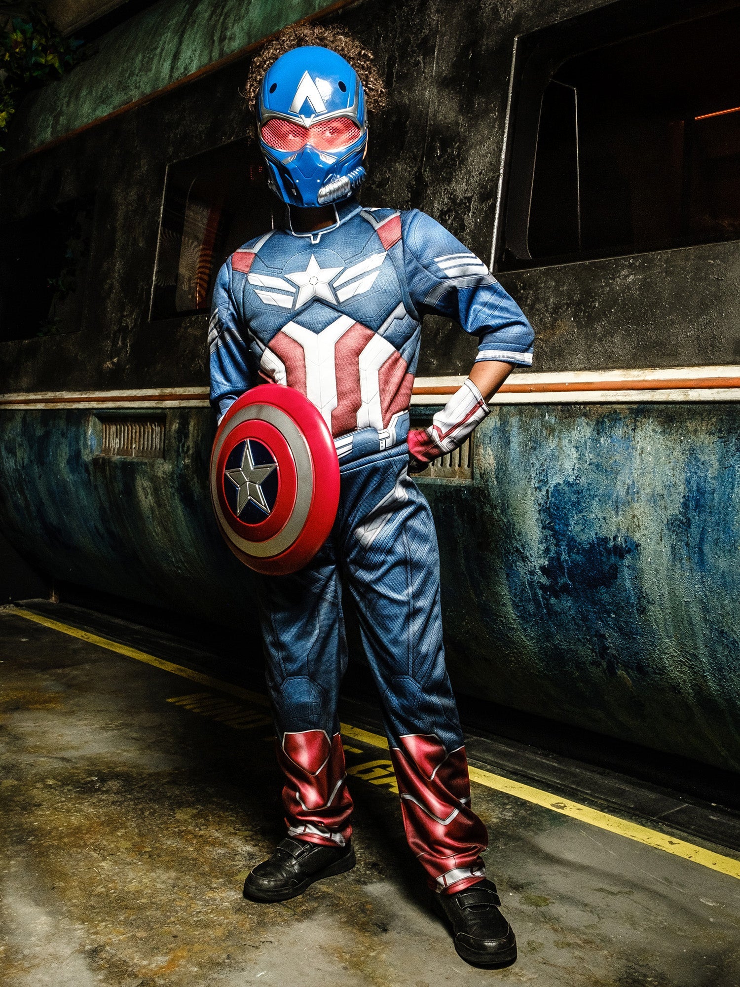 A child in a Captain America costume and mask, posing heroically with one hand on their hip and the other holding a round shield, in front of a grungy, metallic background.