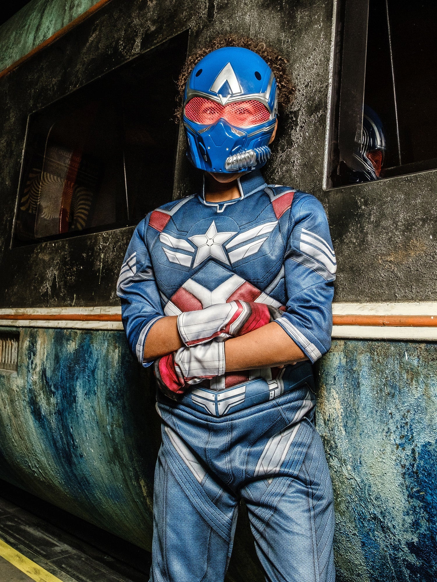 A child in a Captain America costume and mask, standing with arms crossed and a serious expression in front of a grungy, metallic background.