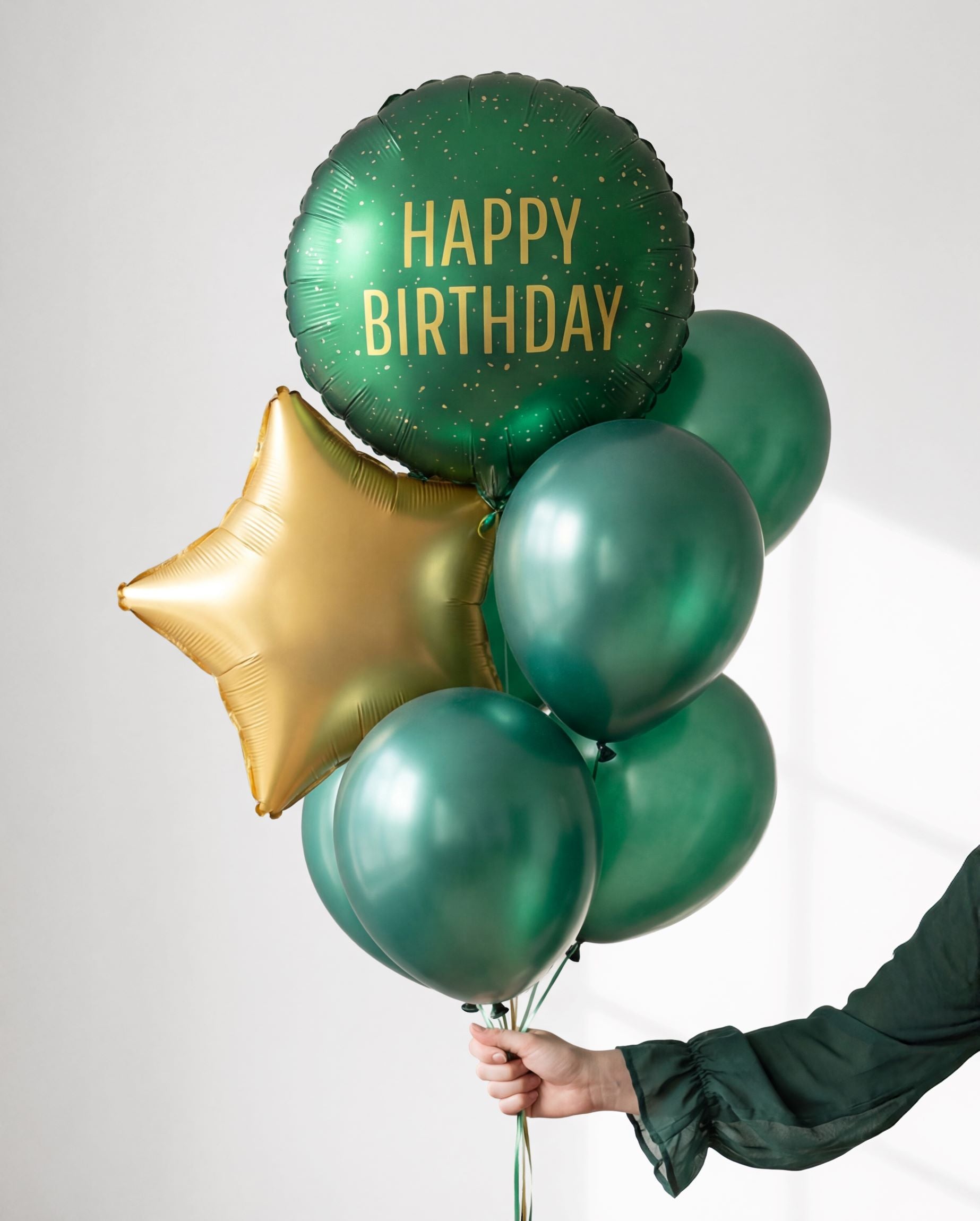 Hand holding a festive balloon bouquet featuring the dark green Happy Birthday foil, a gold star, and matching green latex balloons.