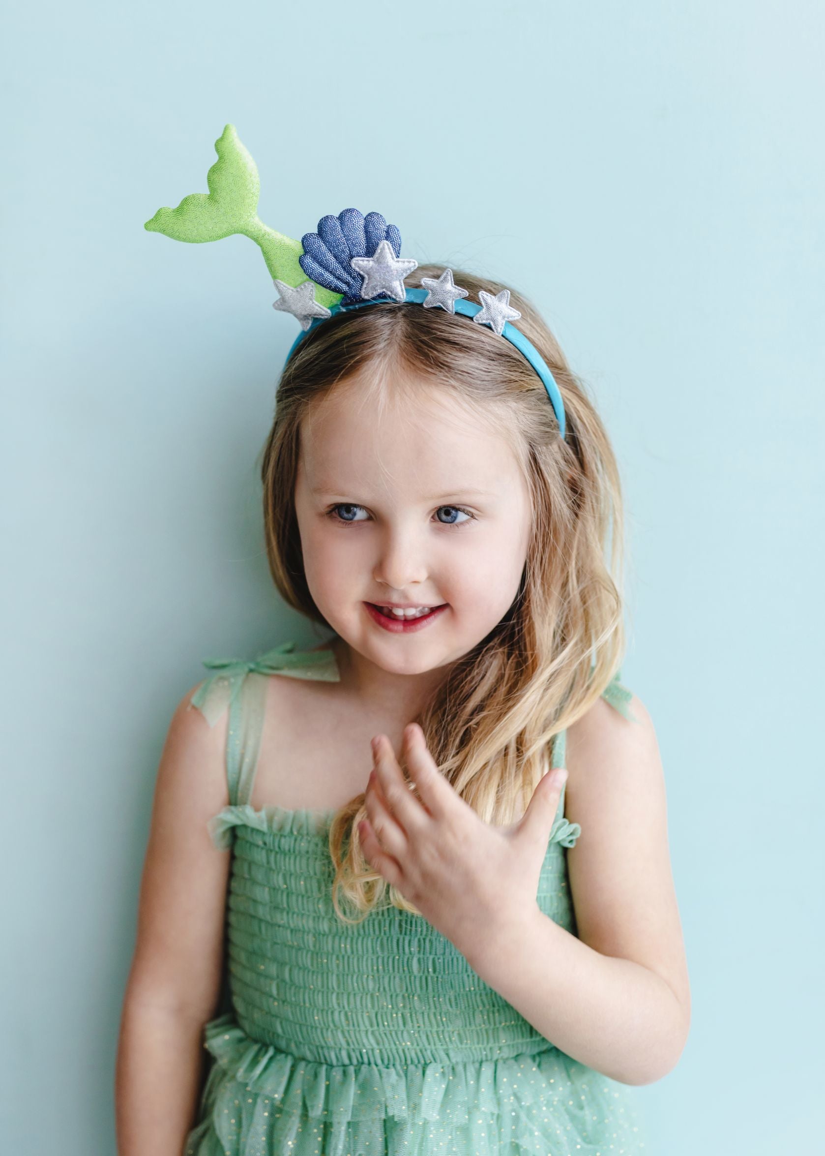 A young child with long, wavy light brown hair smiling and looking to the side, wearing a blue headband adorned with a sparkling green mermaid tail, a blue seashell, and silver glitter starfish, against a plain light blue background.