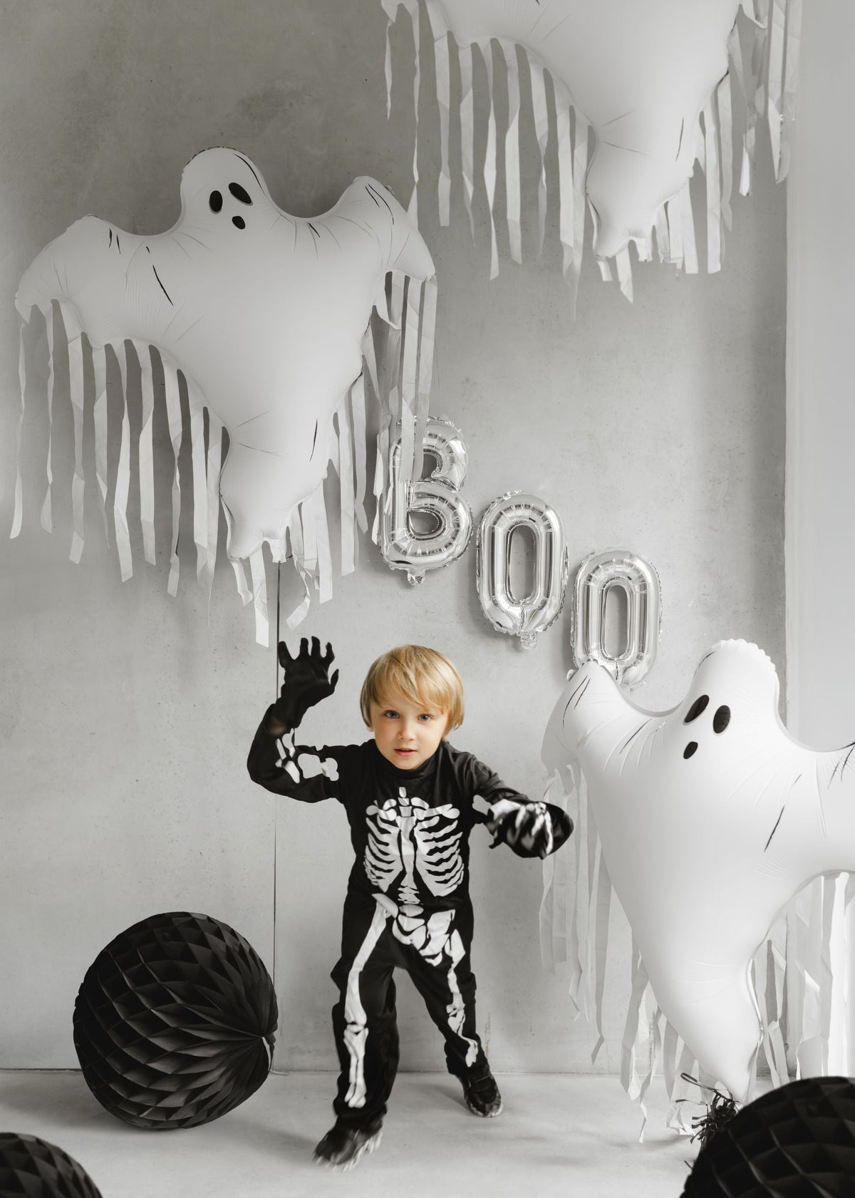 A child dressed in a black skeleton costume, posing with hands raised, in a Halloween-decorated room. Large white foil ghost balloons with tassels hang from the ceiling, along with silver "BOO" letter balloons and black honeycomb balls.