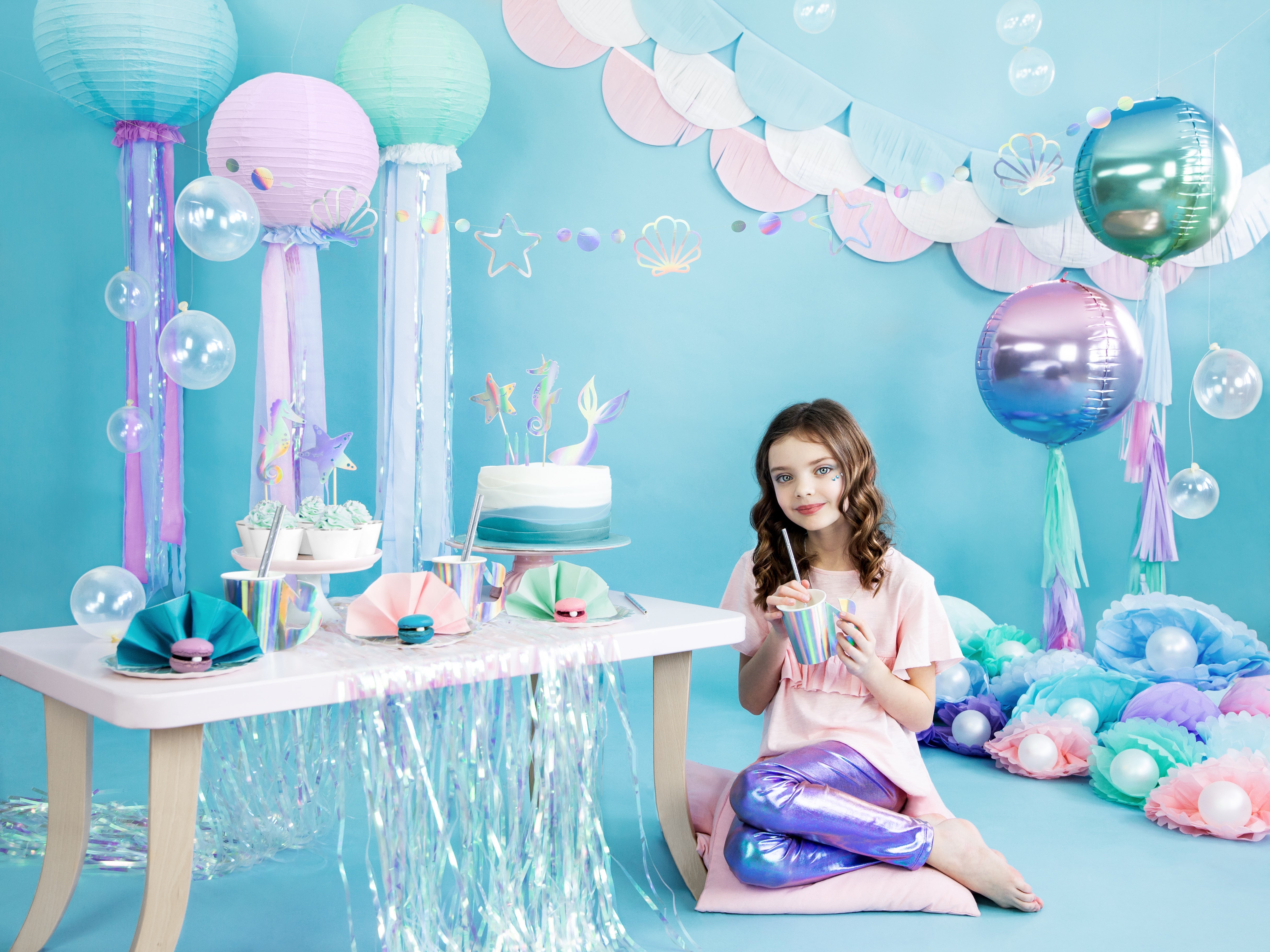 A young person in mermaid-style pants sitting beside a table decorated with mermaid-themed party supplies, including a cake, paper lanterns, balloons, and shimmering fringe, all against a blue background.