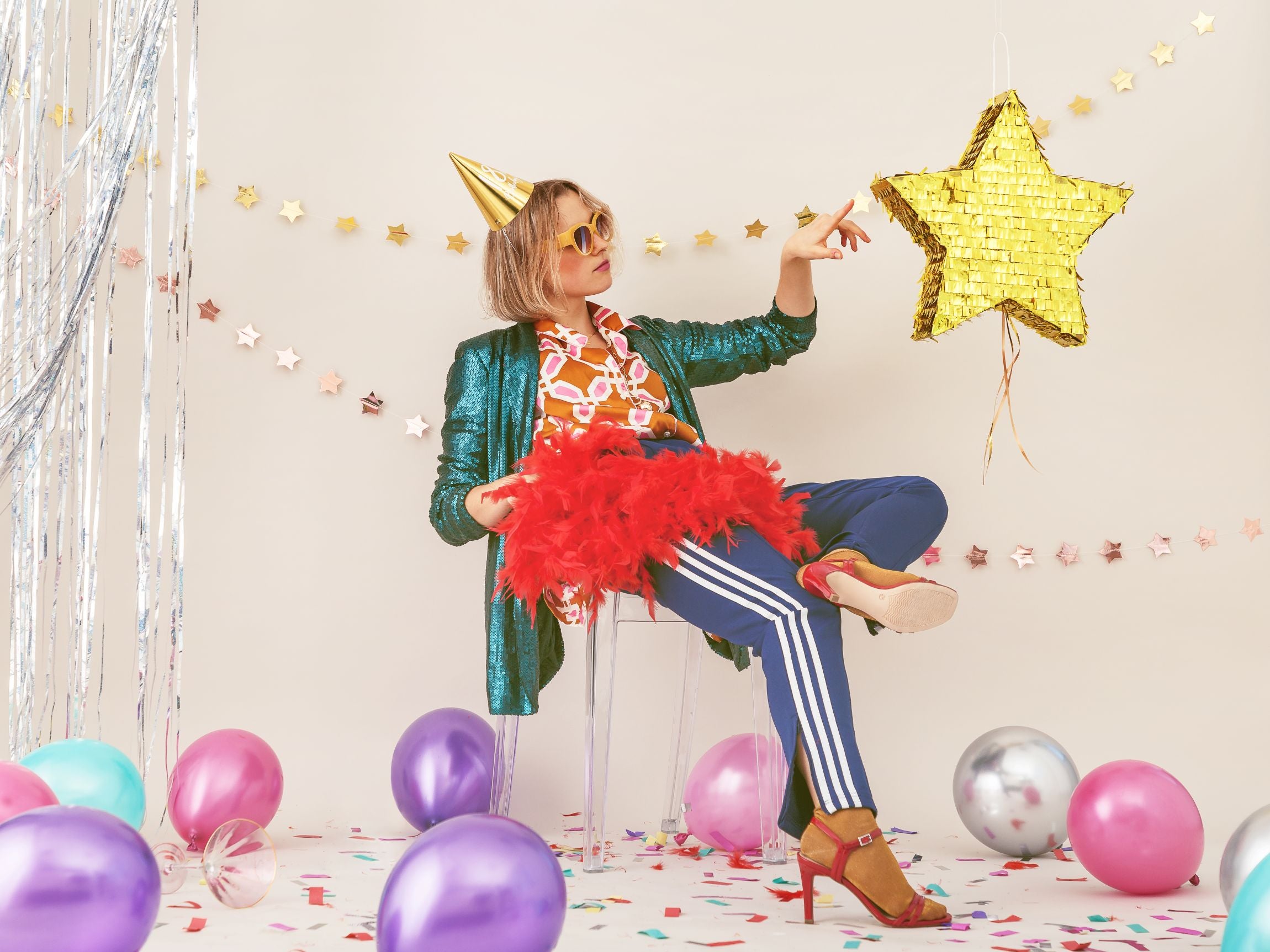 A person wearing a gold party hat and sunglasses, dressed in a green sequin jacket, colorful patterned shirt, and blue striped pants. They are seated, holding a red feather boa, with balloons and streamers around, in a festive party setting