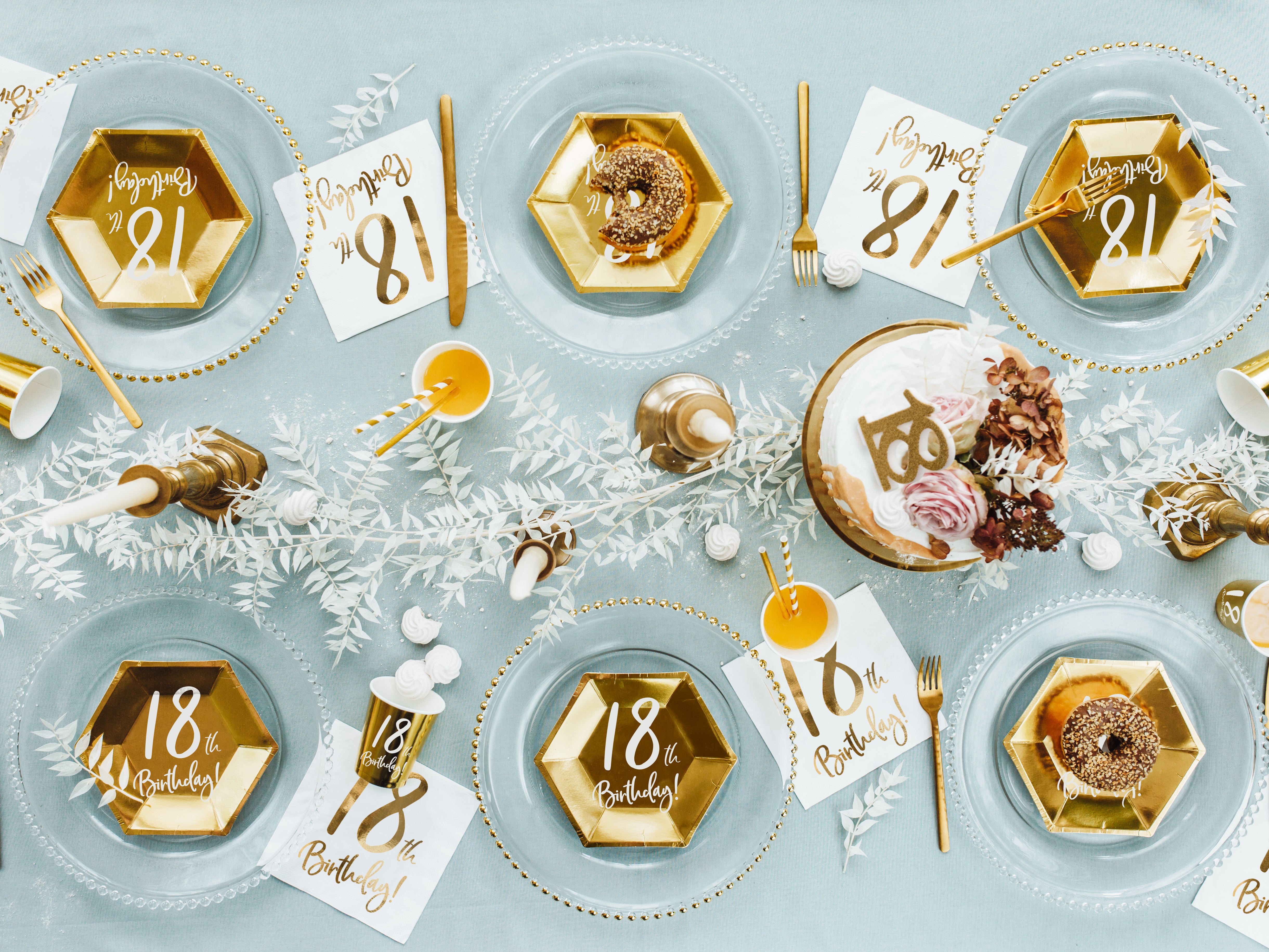 An overhead view of a beautifully set table for an 18th birthday celebration, featuring clear glass charger plates, gold hexagonal paper plates, matching gold cups, gold cutlery, white paper napkins with gold "18th Birthday!" print, and various white decorative elements on a light blue tablecloth.
