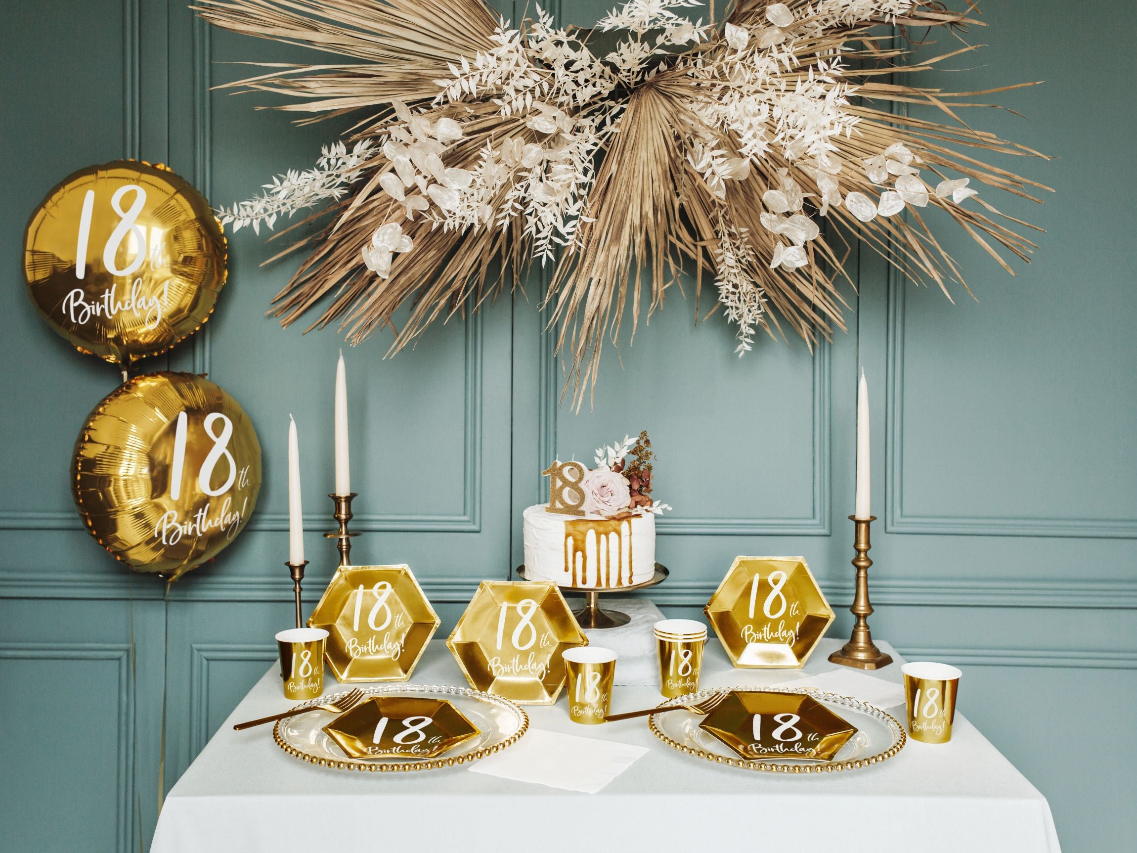 A beautifully decorated table for an 18th birthday celebration, featuring a white tablecloth, gold hexagonal plates, matching cups, tall white candles in gold holders, and a white cake with gold drip and floral decoration. Gold "18th Birthday!" foil balloons and decorative dried palm leaves hang in the background against a teal wall.