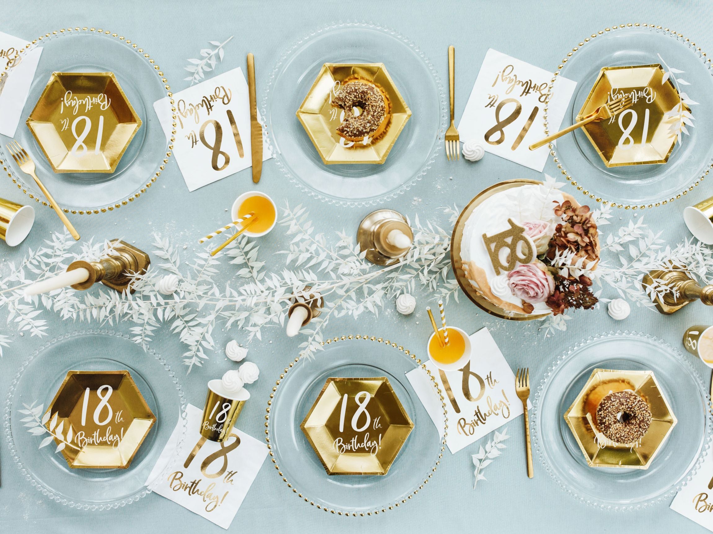 An overhead view of a fully set dining table for an 18th birthday party. The table features clear glass charger plates, topped with gold hexagonal paper plates printed with "18th Birthday!", gold cutlery, matching gold cups, small desserts, a birthday cake, and scattered white decorative foliage, all on a light blue tablecloth.