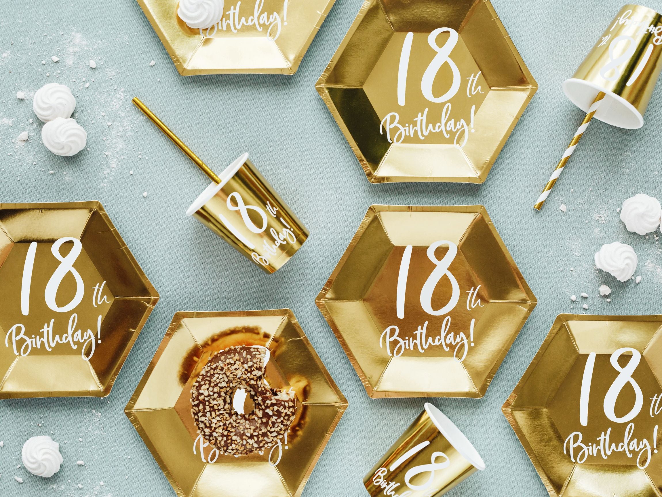 An overhead flat lay of multiple hexagonal metallic gold paper plates, each printed with "18th Birthday!" in white. Some plates have partial donuts with sprinkles and small white meringues scattered around, along with matching gold cups and a gold striped straw, all on a light blue background.
