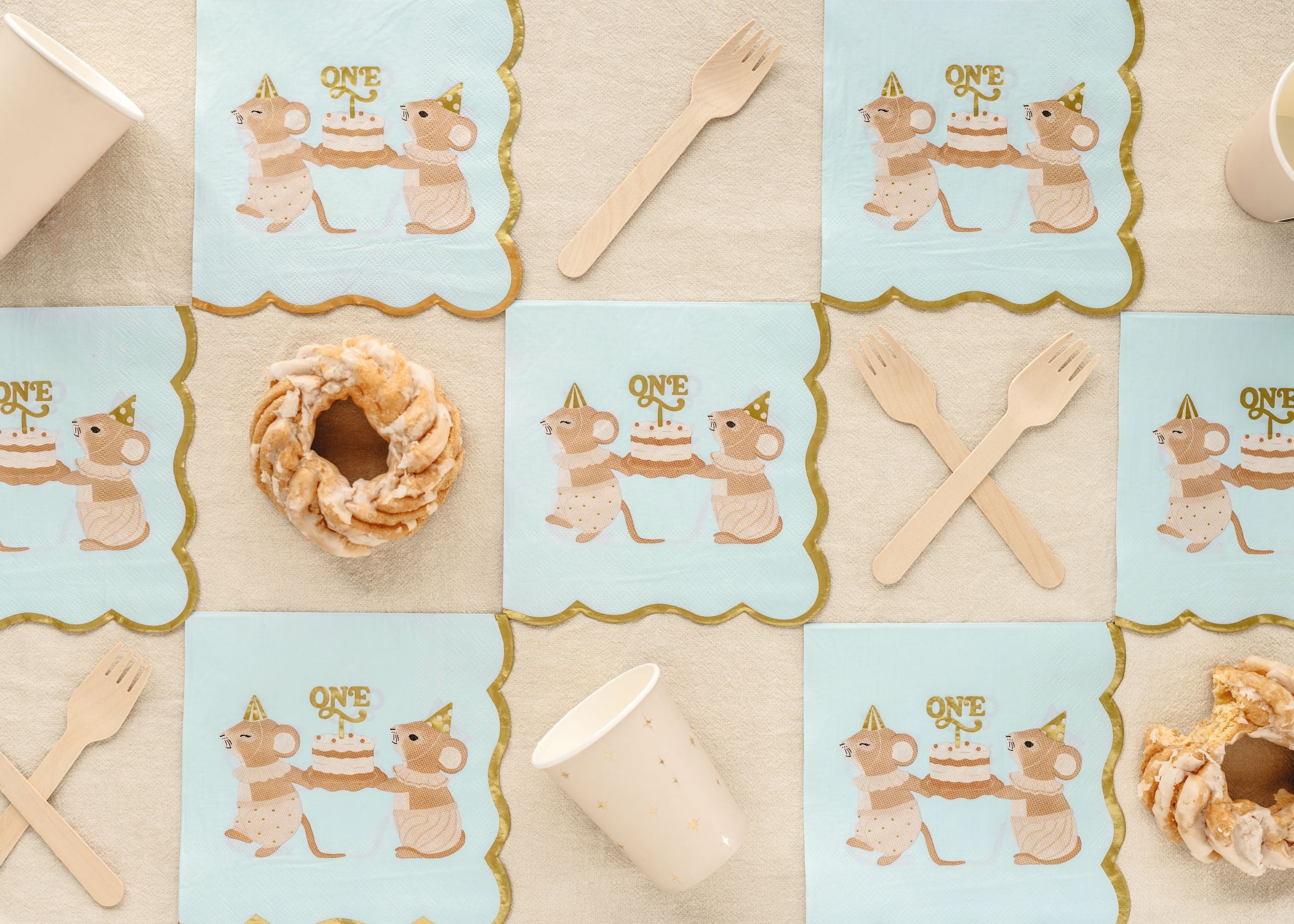 A top-down view of blue 1st birthday napkins with a mice and cake design, styled with eco-friendly wooden cutlery and a sweet pastry.