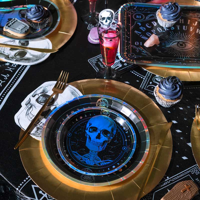 A styled Halloween table setting featuring gold paper plates with blue skull and occult-themed designs, black patterned tablecloths, cupcakes, and a pink drink in a glass.
