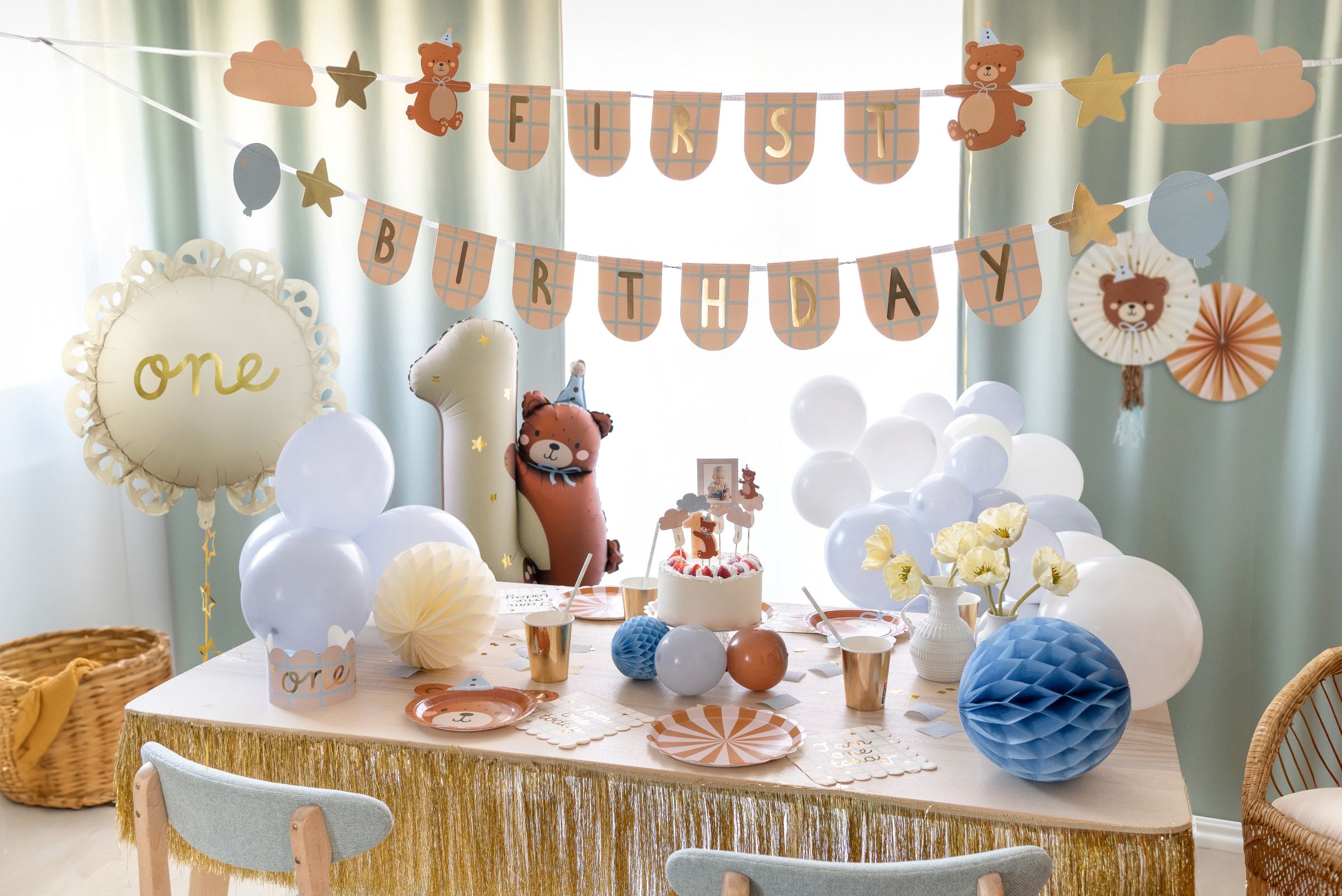 A close-up of a charming teddy bear themed party display, showing a large decorative paper rosette with a teddy bear face, smaller gold star foil balloons, and a "BIRTHDAY" banner with plaid patterns and teddy bear cutouts, all set against a white curtain.