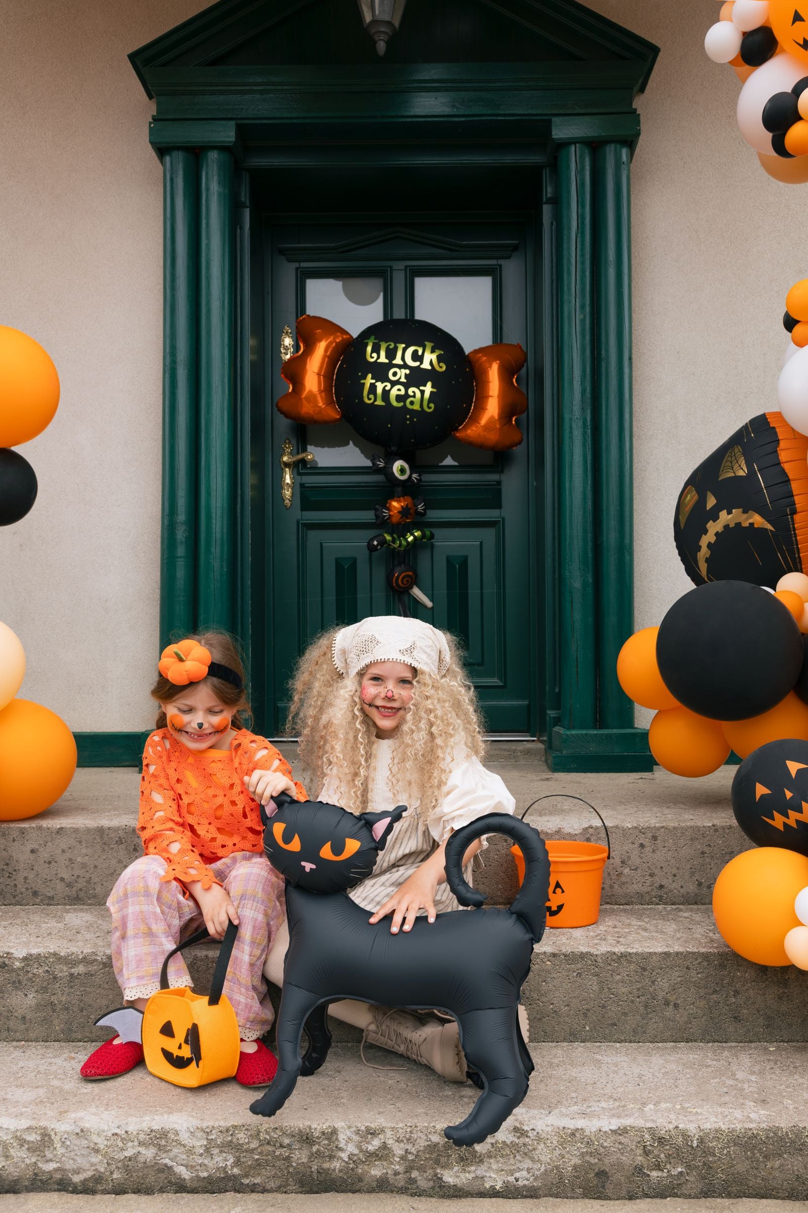 A woman and child in Halloween costumes sit on stone steps in front of a dark green door decorated for Halloween, with a black cat-shaped balloon, orange pumpkins, and black-and-orange balloon clusters around them.