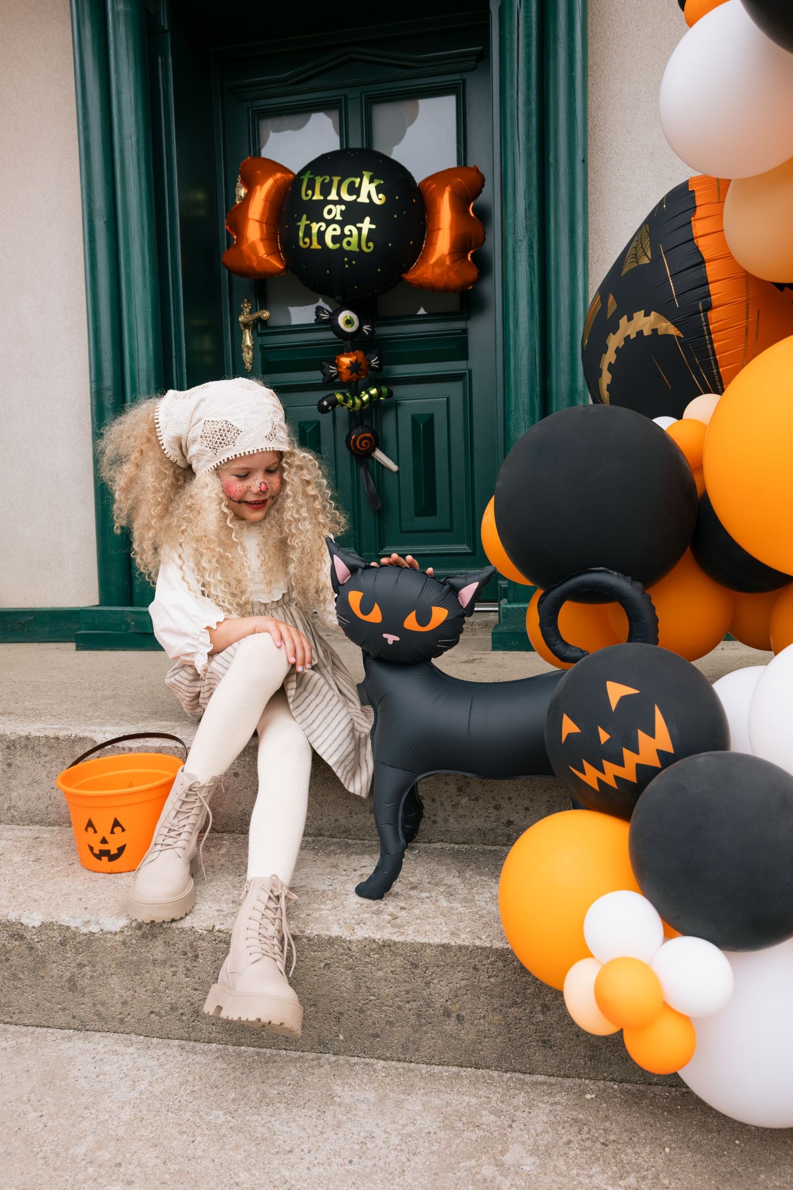 Young girl with curly blonde hair and Halloween face paint sitting on stone steps, petting a standing black cat foil balloon.