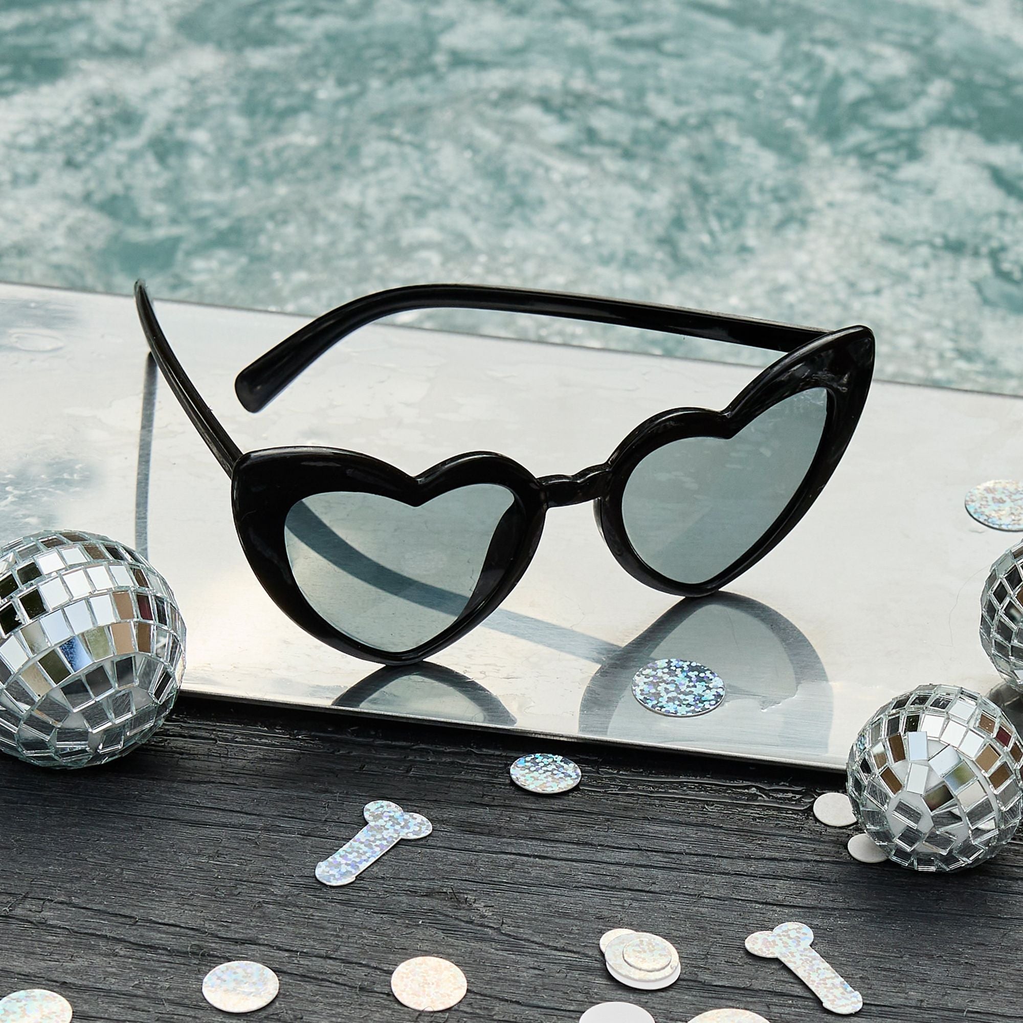 Black heart-shaped sunglasses resting on a silver surface by a pool, surrounded by mini disco balls and silver confetti.