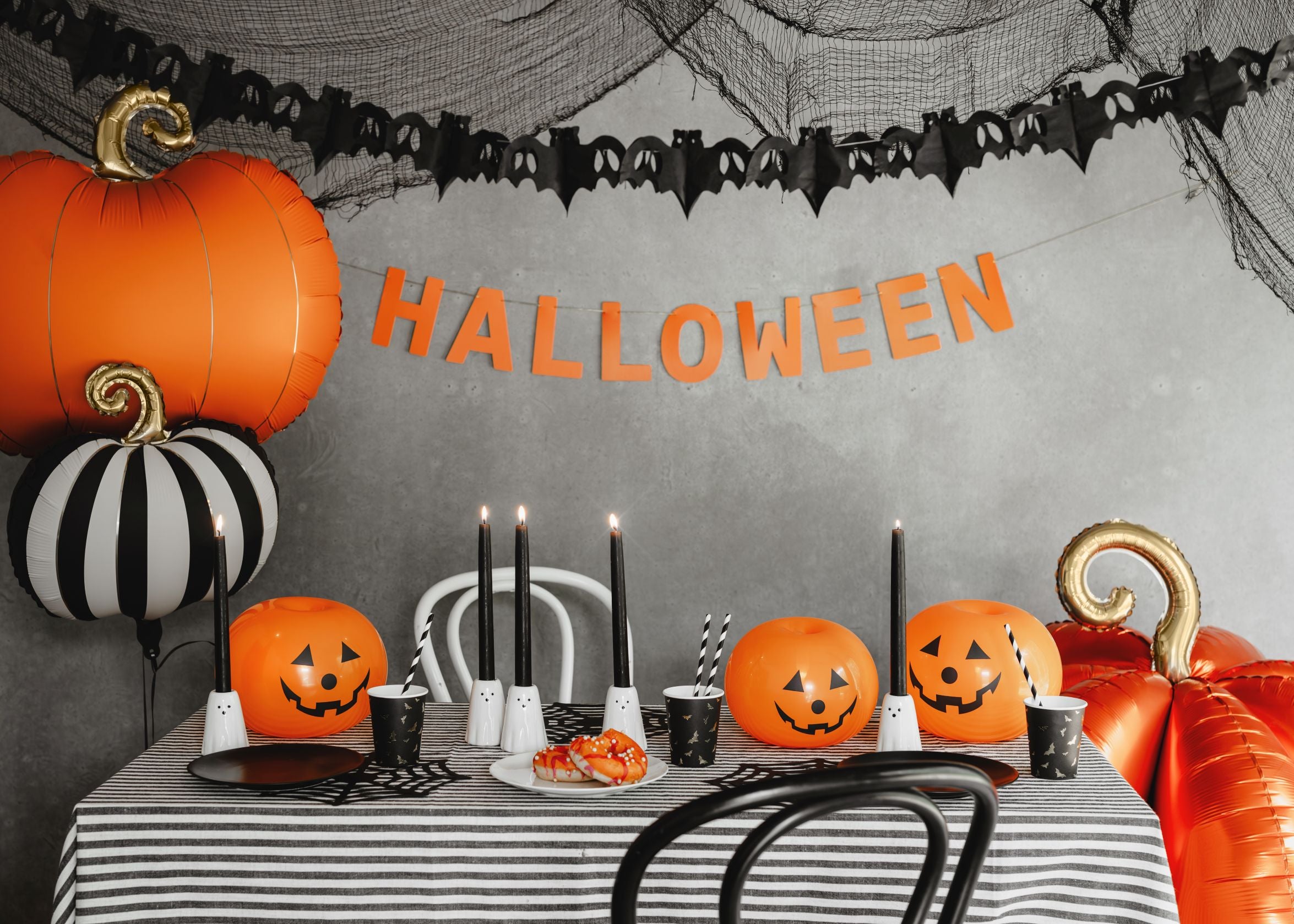 A stylish Halloween party table setup against a grey wall draped with black cobweb netting and a bat banner. The table is covered with a black and white striped tablecloth and decorated with black candles, jack-o'-lantern shaped cups, and features a large orange pumpkin foil balloon and a matching black and white striped pumpkin foil balloon.
