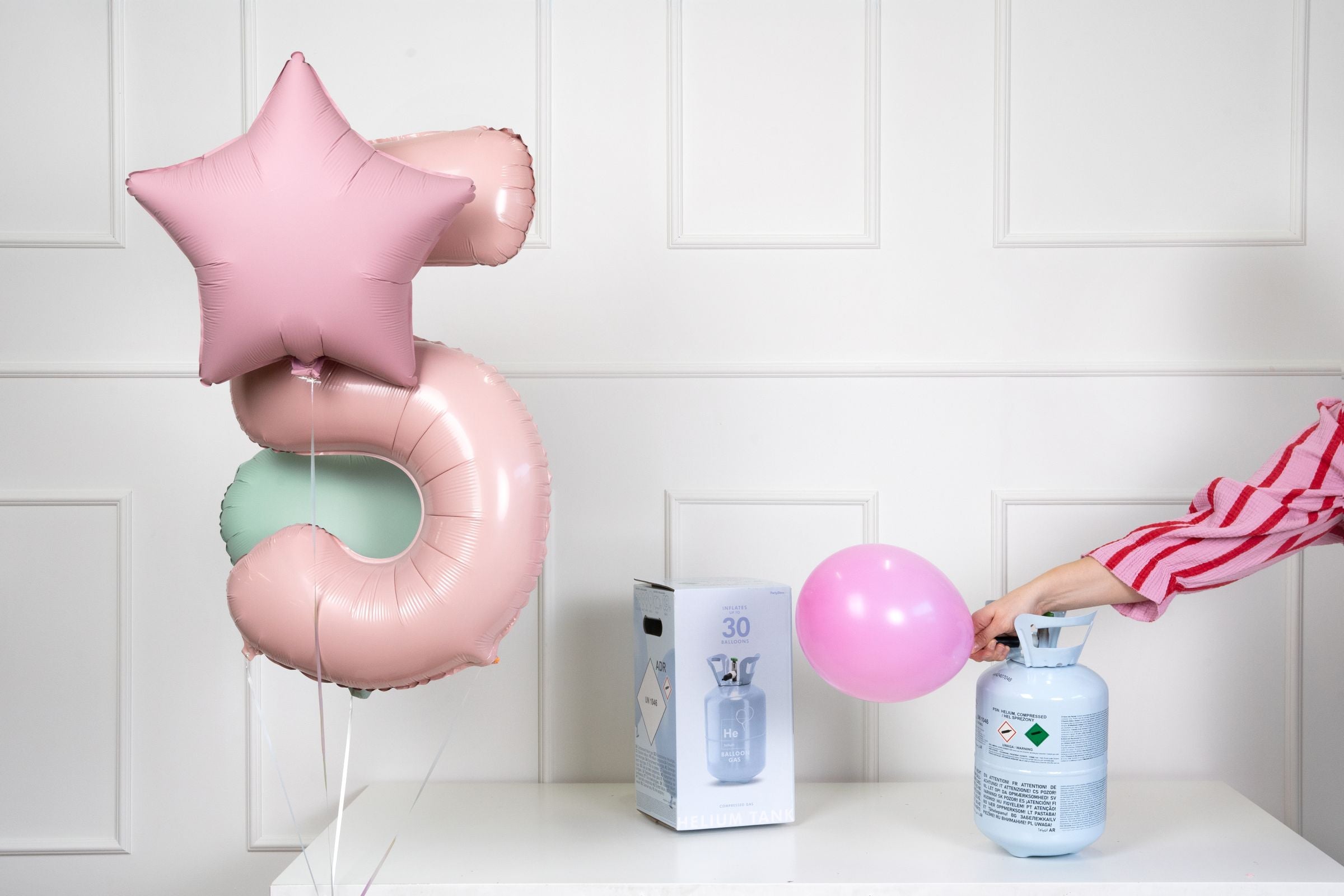 A light blue helium balloon tank and its product box on a white surface, next to a pink number "5" foil balloon and a pink star foil balloon. A hand is seen inflating a pink latex balloon from the canister, against a white paneled wall.